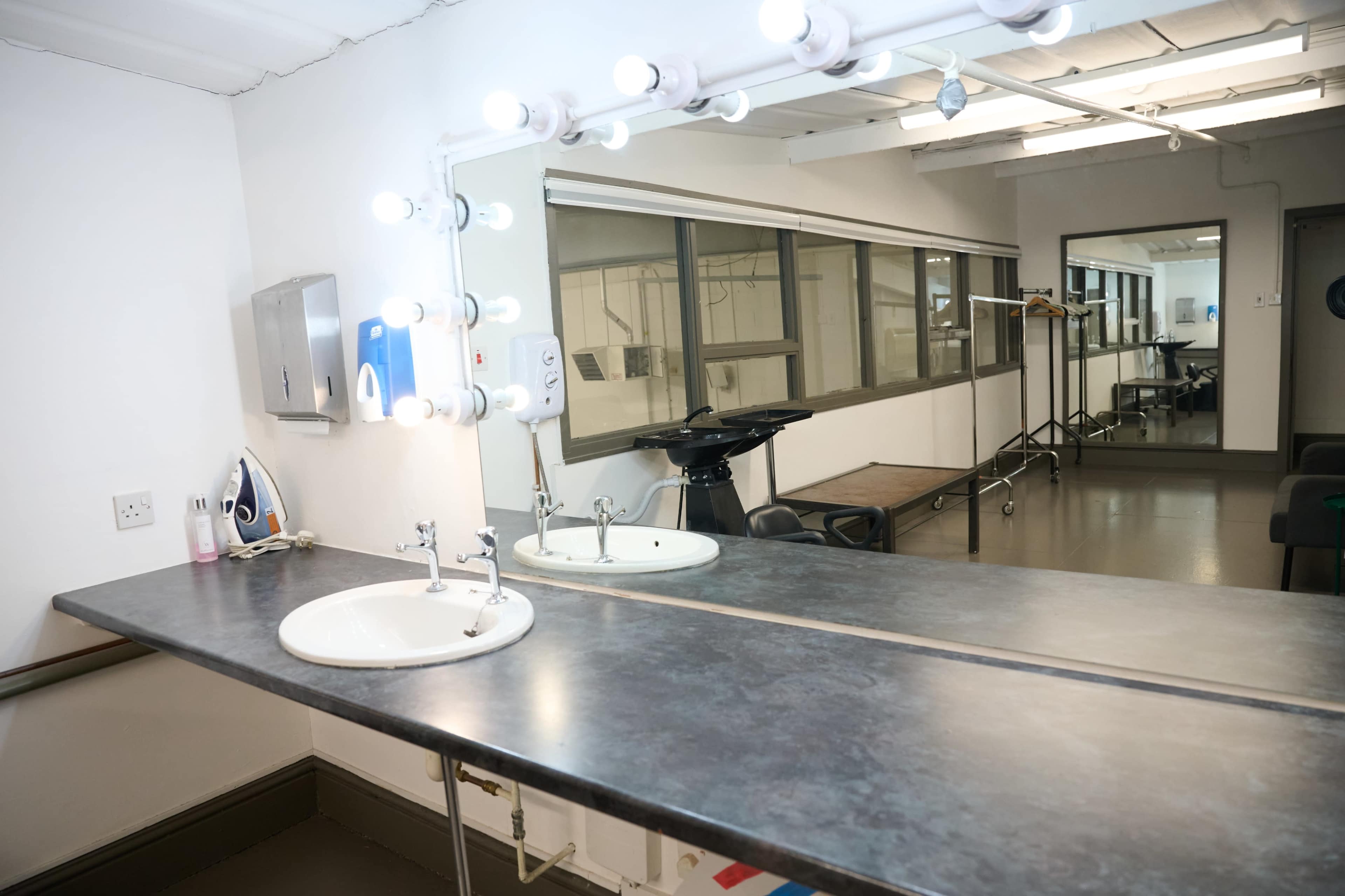 The image shows a modern vanity area with a long countertop featuring two sinks, illuminated by a row of vertical light bulbs, and a reflection of a room with equipment in the background.