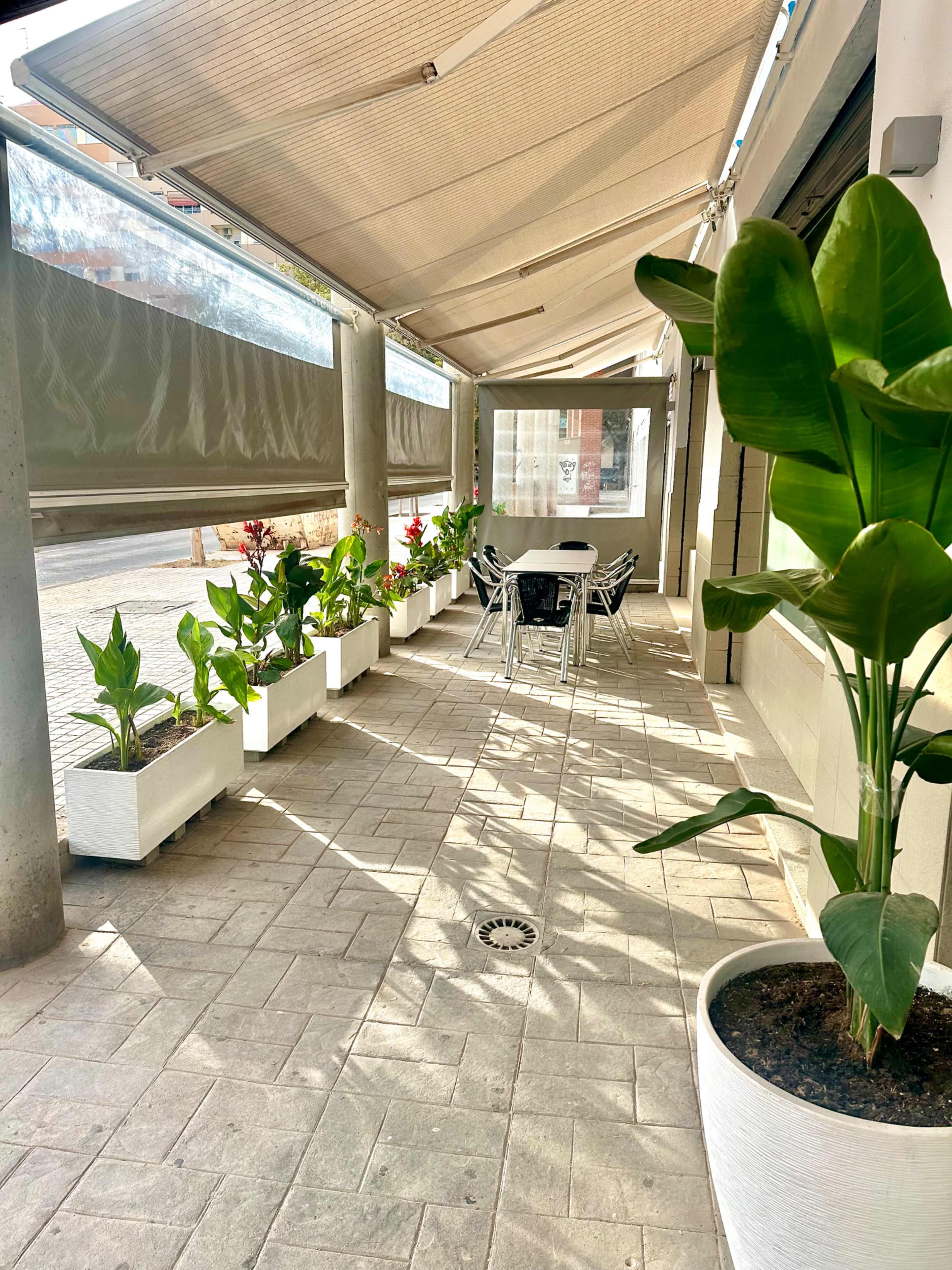 A covered outdoor seating area features a row of potted plants lining the walkway beside a table and chairs.