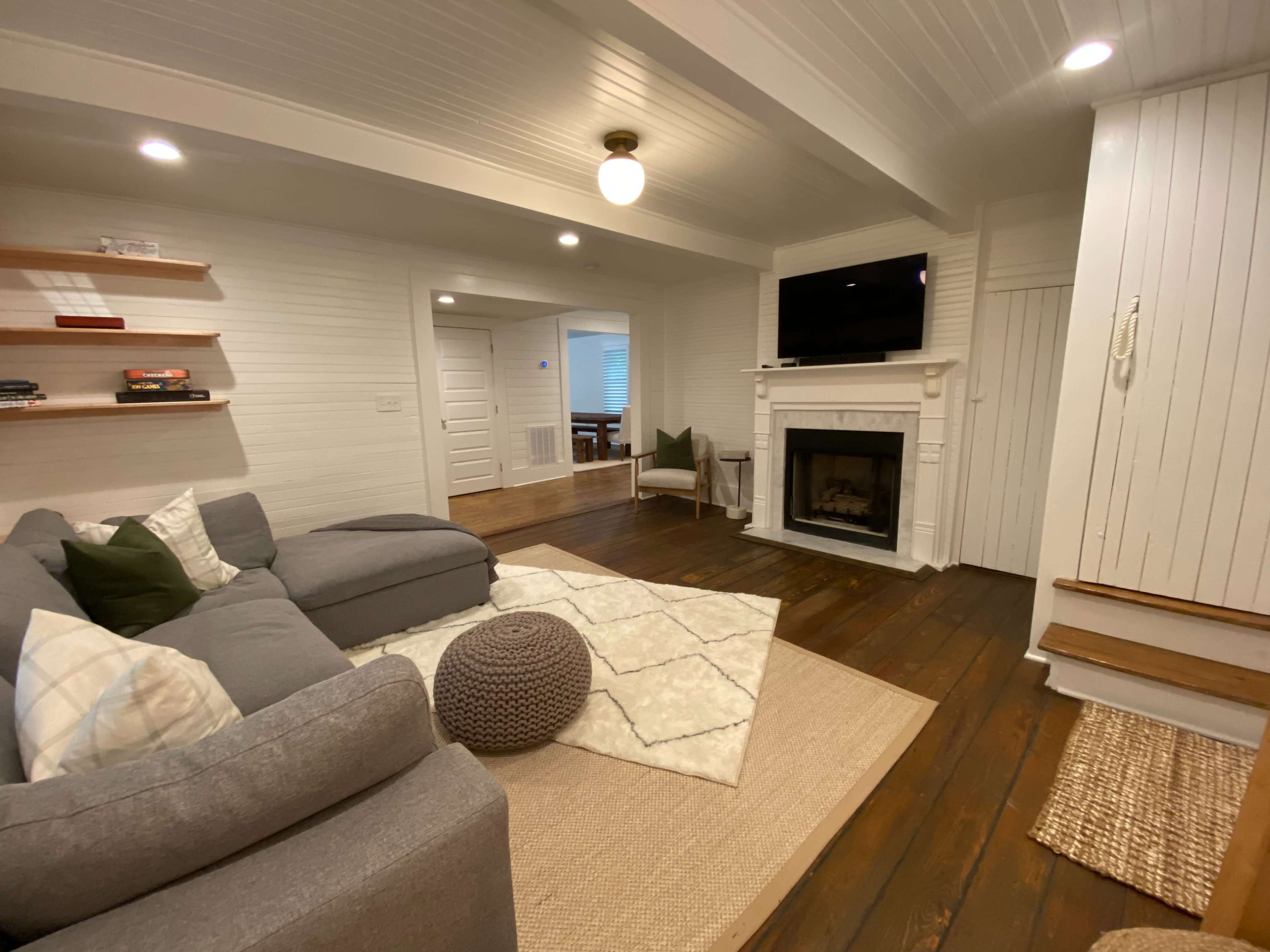 A cozy living room features a gray sectional sofa, a white fireplace with a TV mounted above it, and wooden floorboards.
