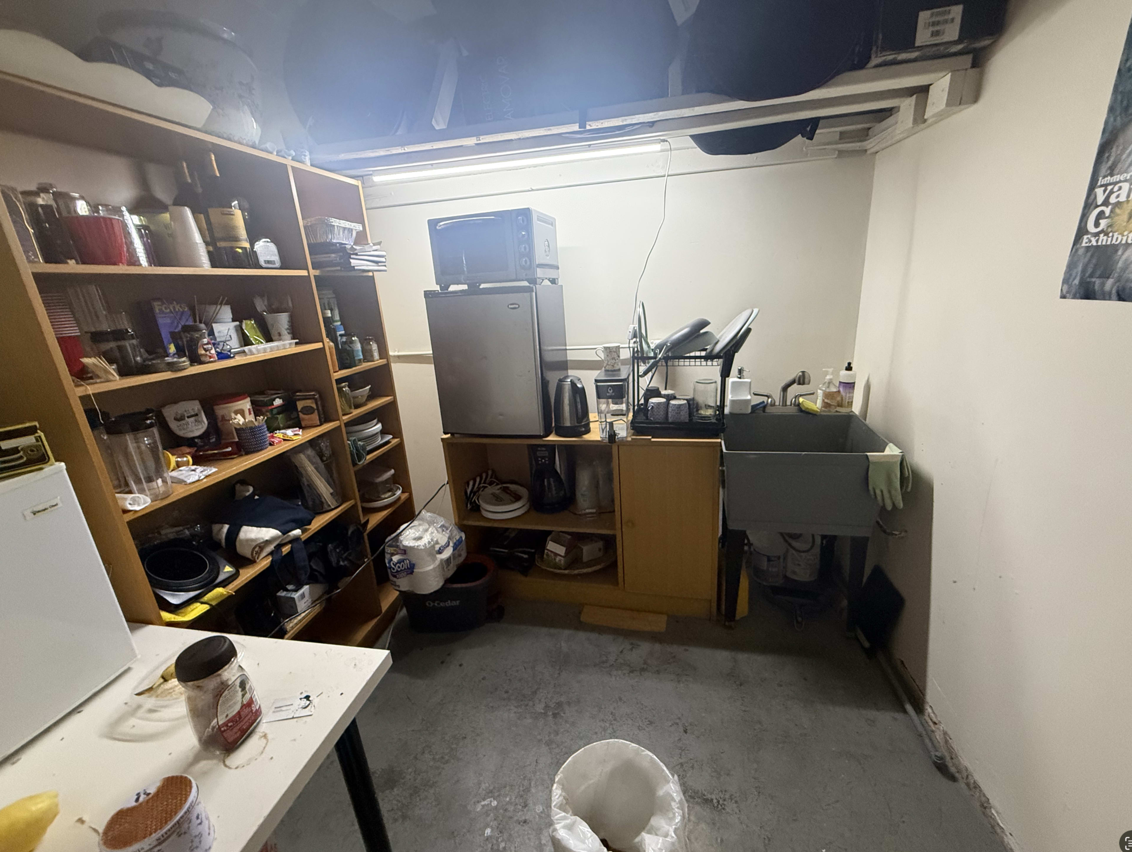 The image shows a small, cluttered kitchen area with shelves filled with various kitchen items, a microwave, and a sink on a counter.