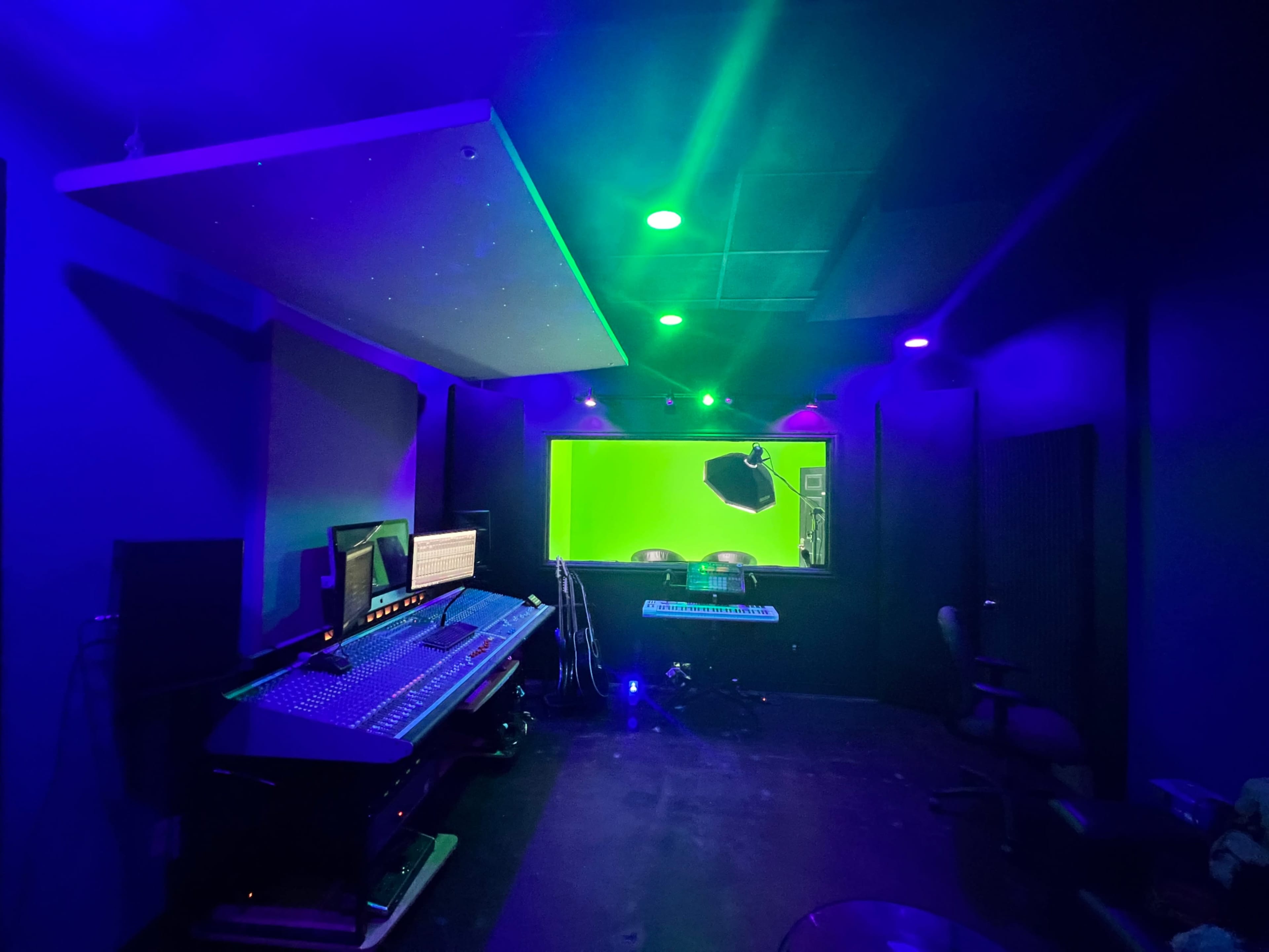 A dimly lit recording studio featuring a sound mixing console, multiple screens, and a green screen setup.