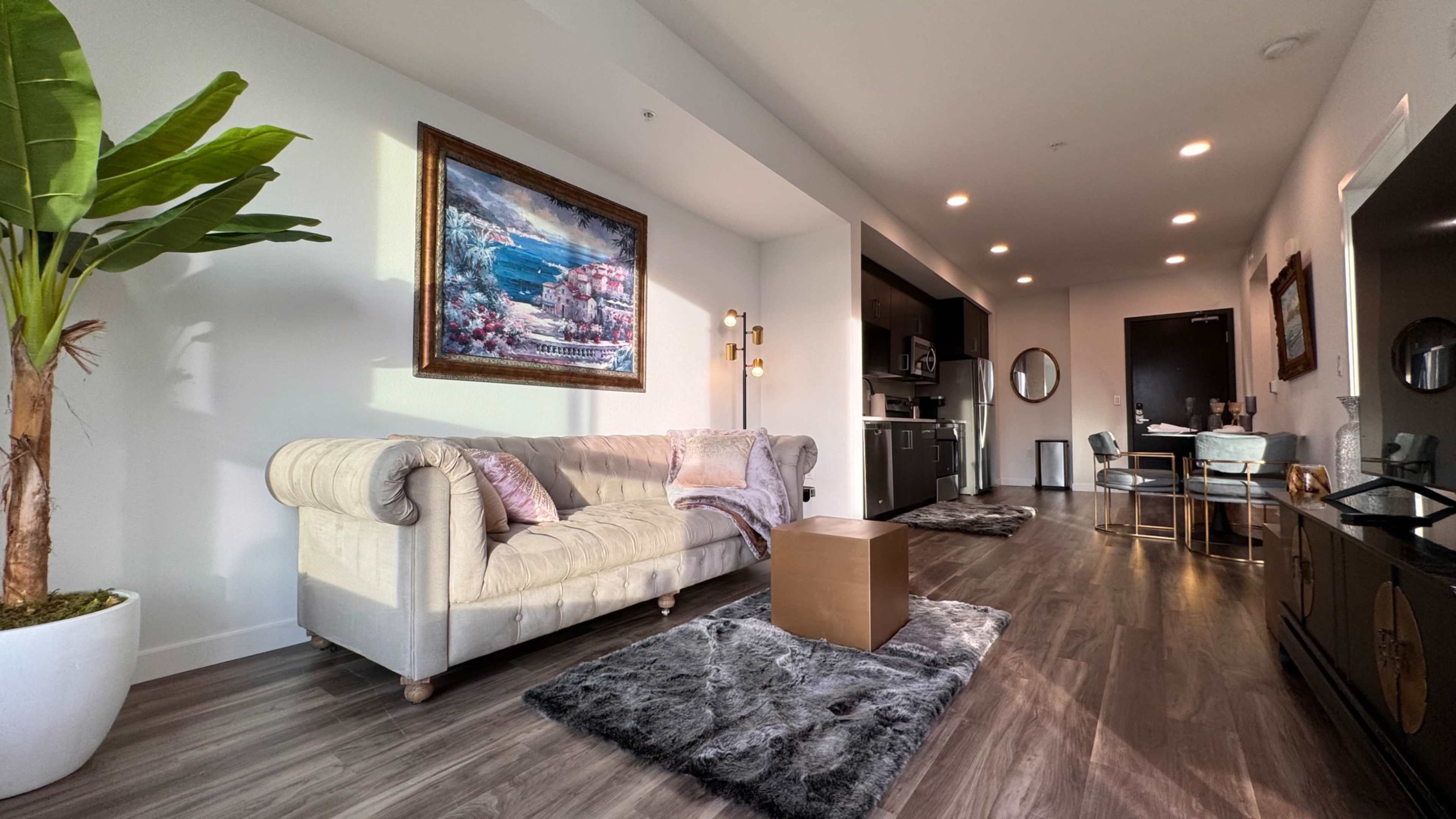 A modern living area featuring a beige sofa, a decorative painting on the wall, a dining area in the background, and a potted plant.