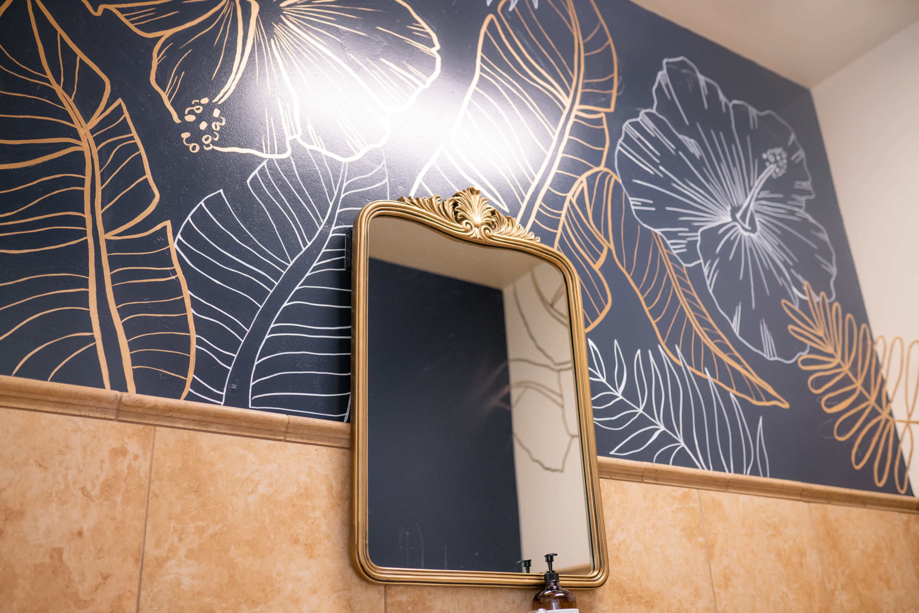A decorative bathroom wall featuring a floral and leaf pattern, with a gold-framed mirror positioned in front of it.