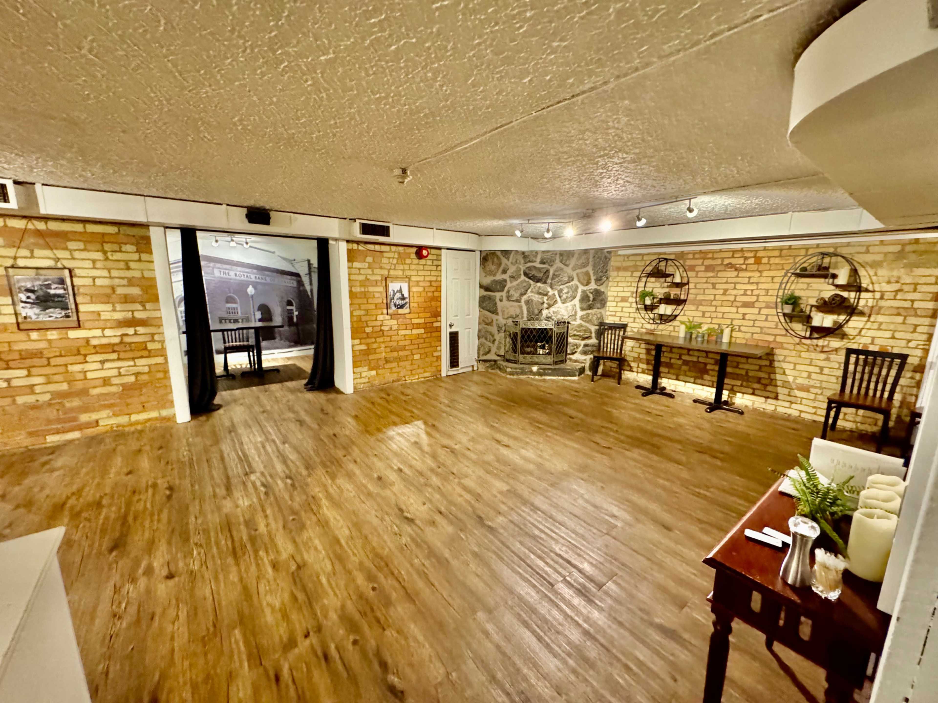 A spacious, empty room with wooden flooring, exposed brick walls, and a stone fireplace, featuring tables and chairs set against one wall.
