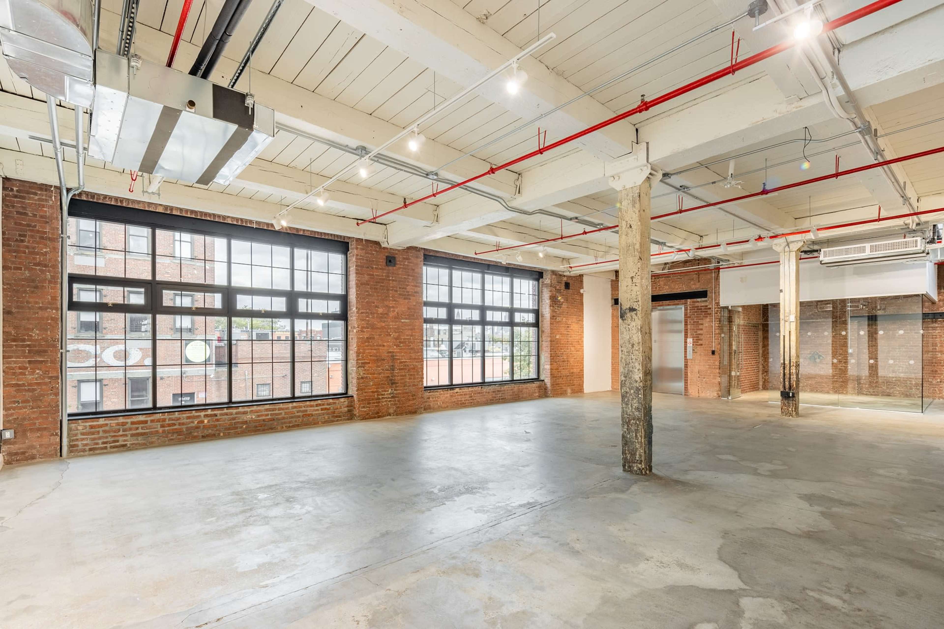 An open, industrial-style space with large windows, exposed brick walls, and concrete flooring.