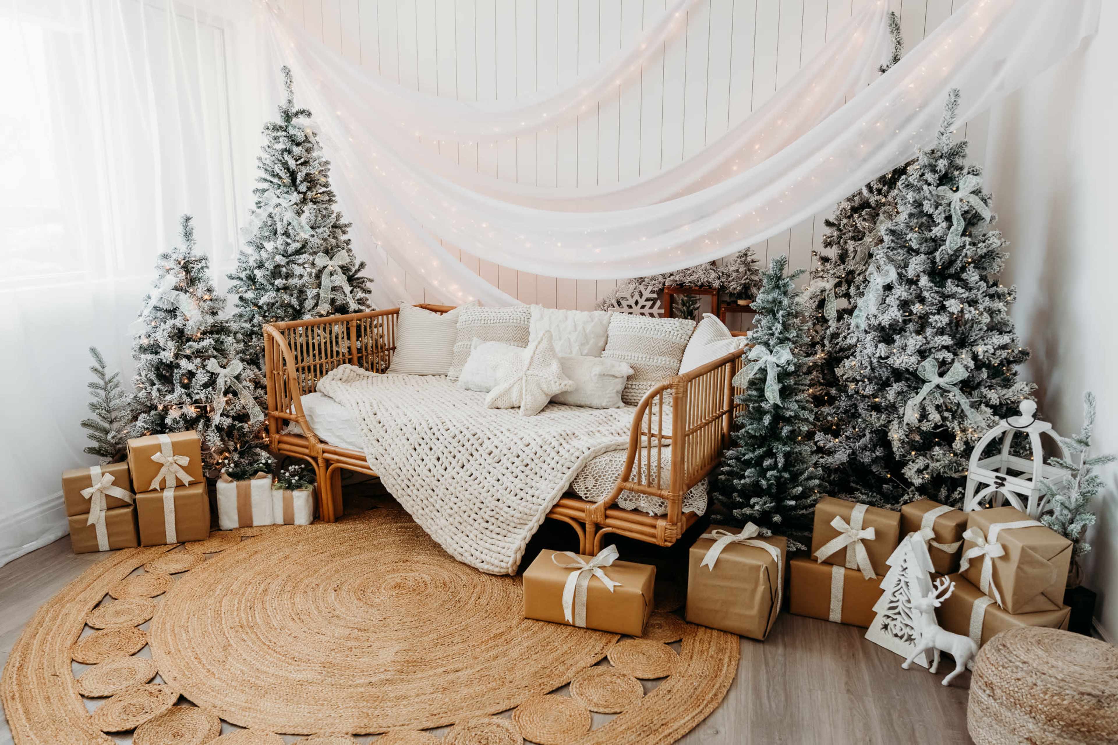 The image shows a cozy indoor scene featuring a rattan daybed adorned with pillows, surrounded by decorated Christmas trees and neatly arranged gift boxes.