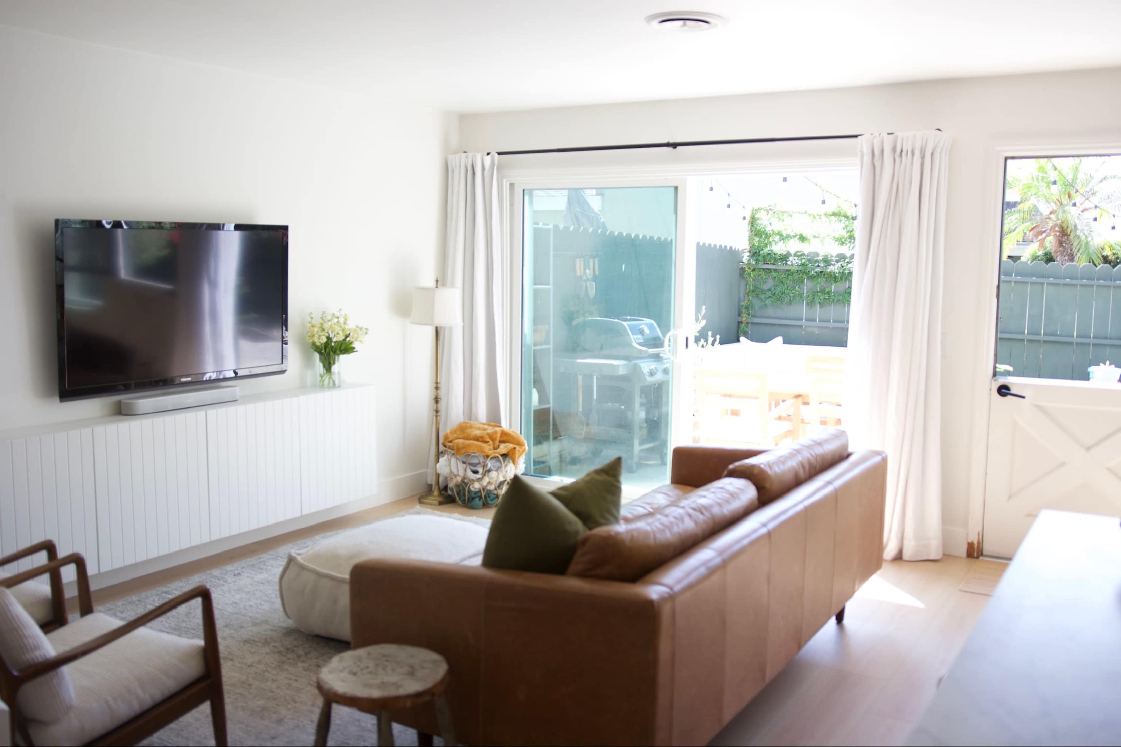 A living room features a leather sofa, a television mounted on the wall, and a door leading to an outdoor area with a dining table.
