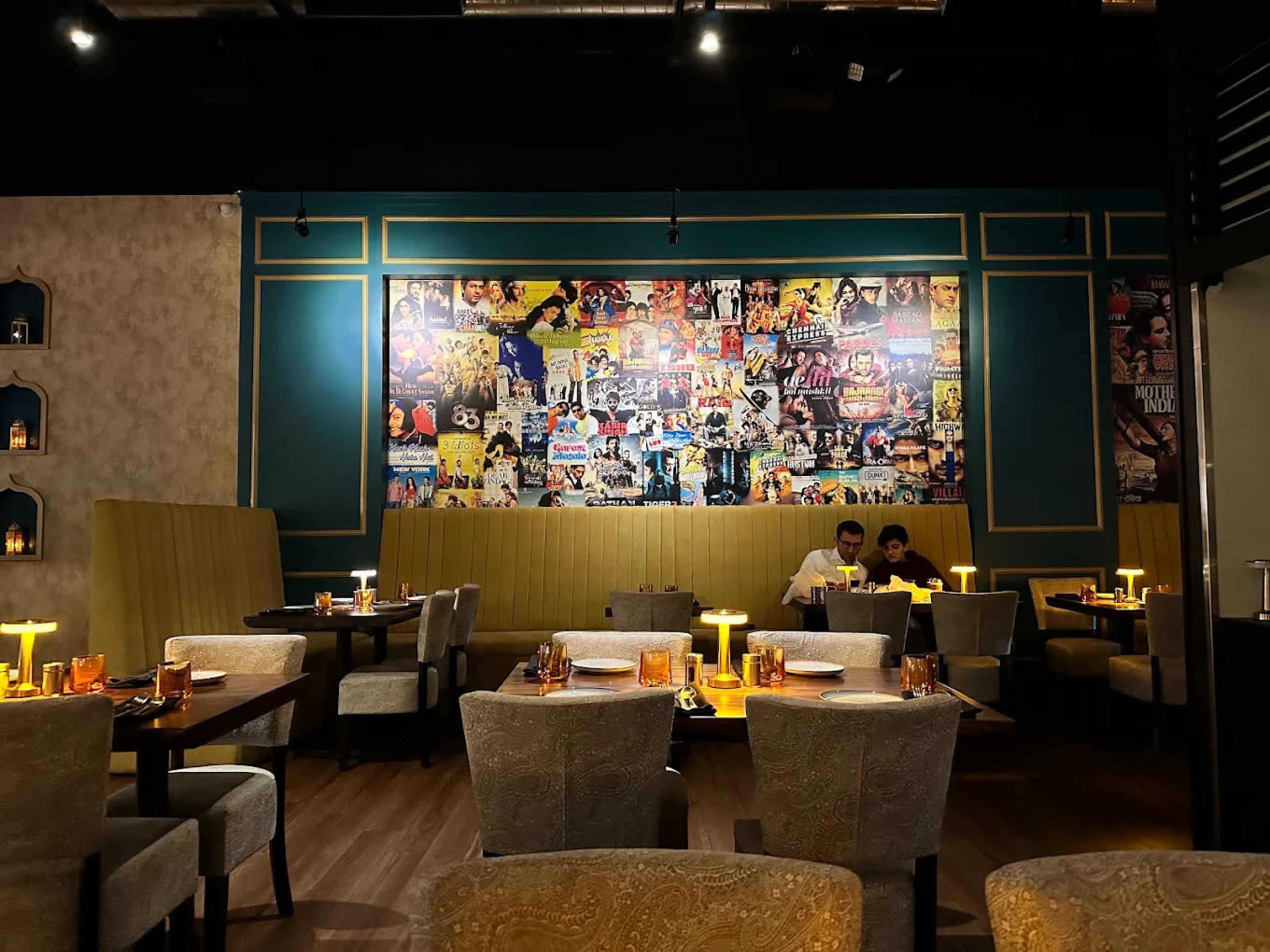 The image shows a restaurant interior with beige chairs and tables, featuring a colorful wall collage made up of movie posters.