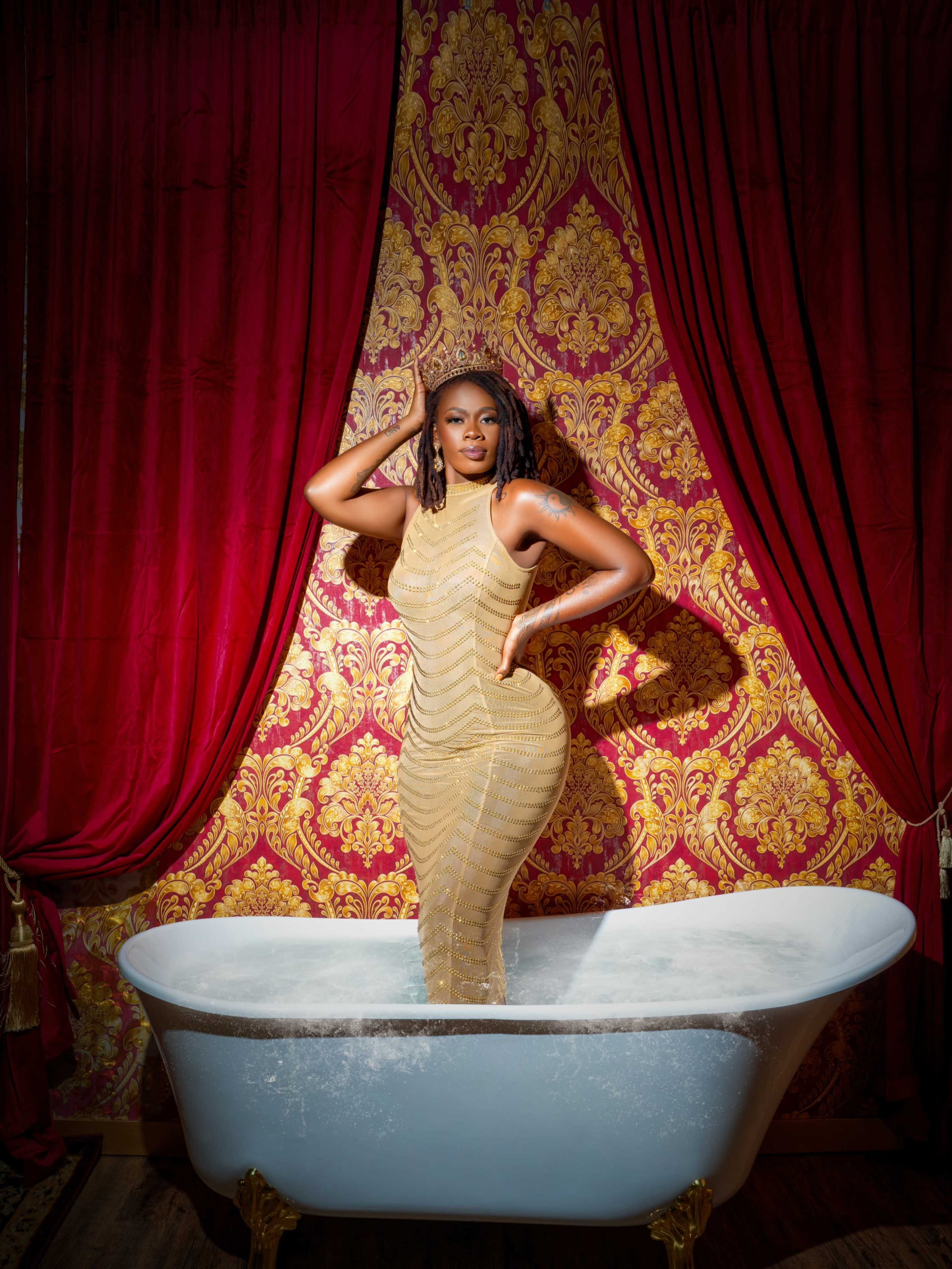 A person stands in a clawfoot bathtub filled with water, framed by red curtains and a patterned golden wall.