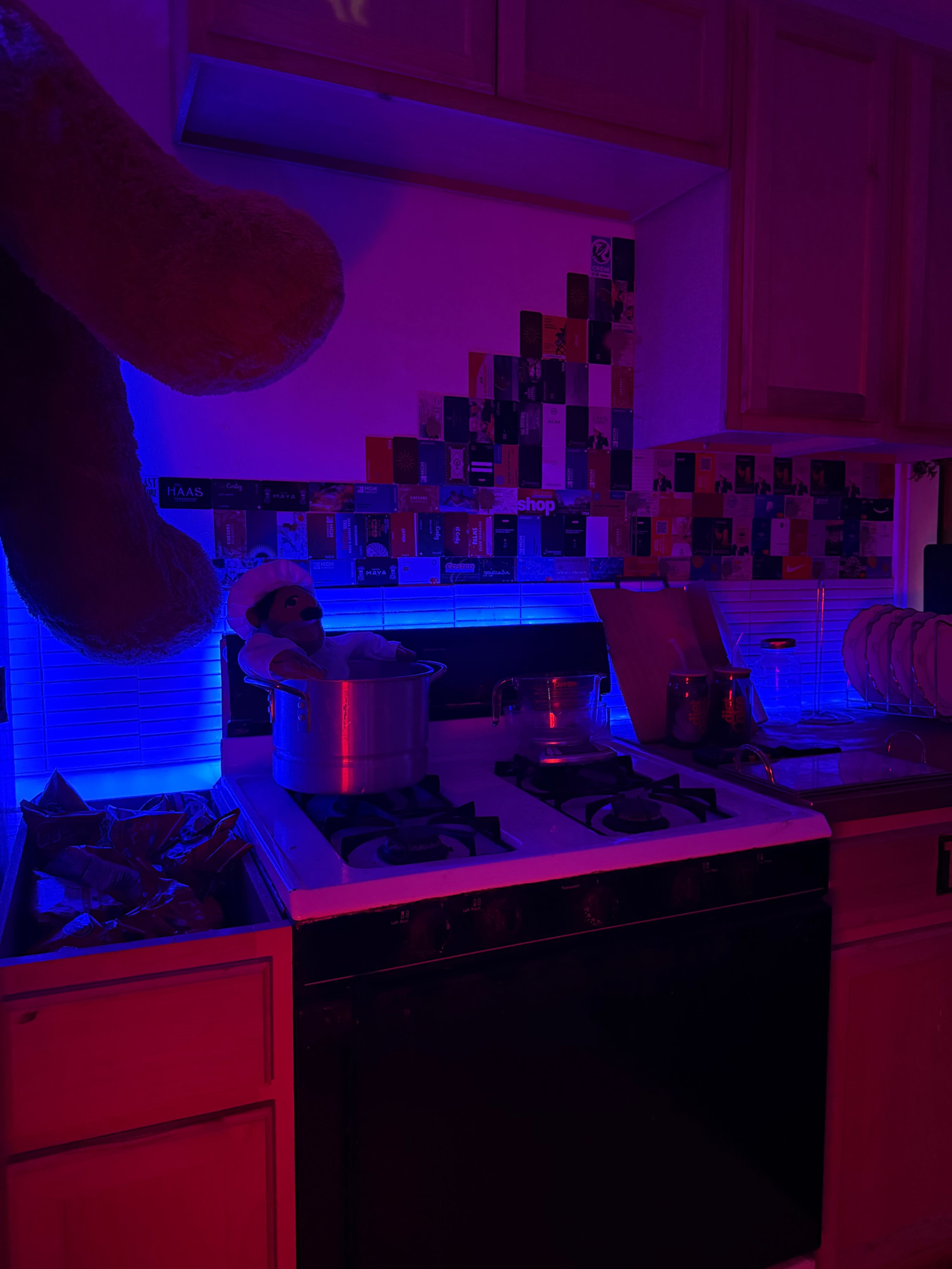 The image shows a dimly lit kitchen with a pot on the stove, colorful wall decorations, and a large teddy bear hanging from the ceiling.
