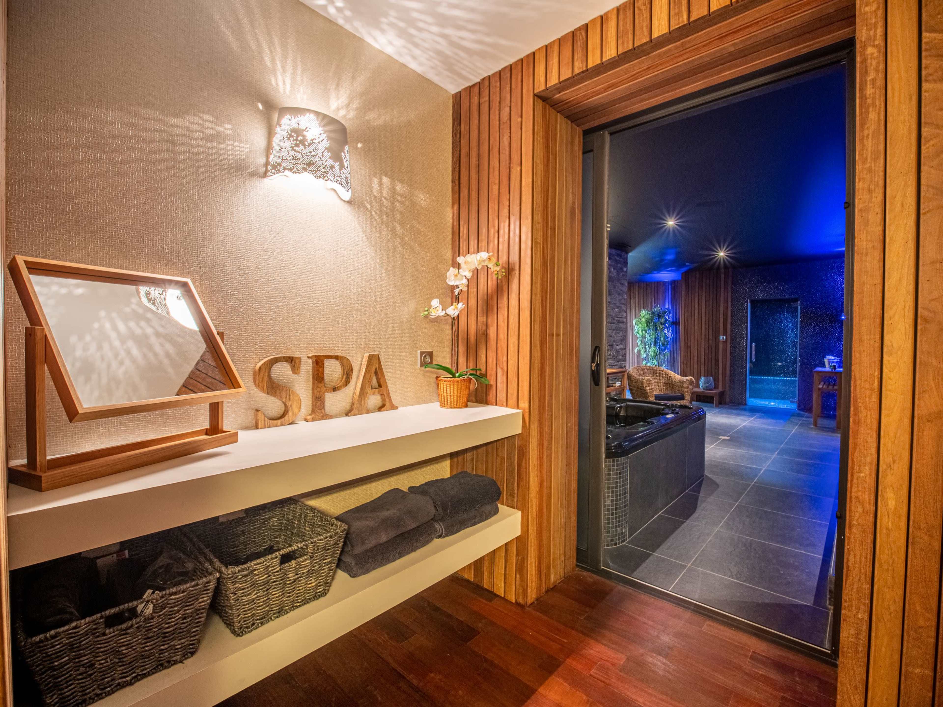 The image shows a spa entryway featuring a wooden door, wall decor displaying the word "SPA," and neatly arranged towels on a shelf alongside wicker baskets.