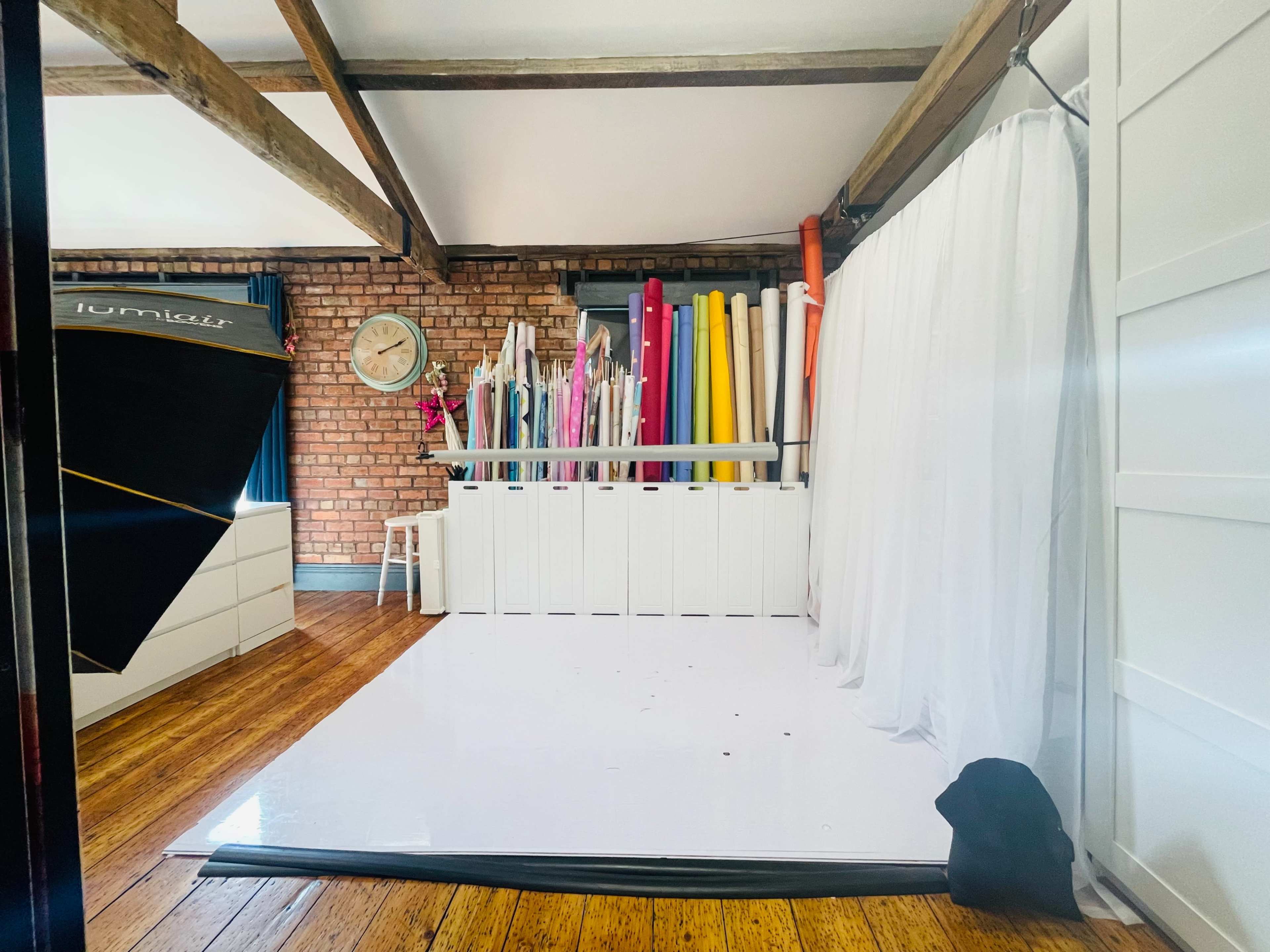 A photography studio with a white floor, a backdrop curtain, and a wall lined with colorful rolls of paper against a brick background.