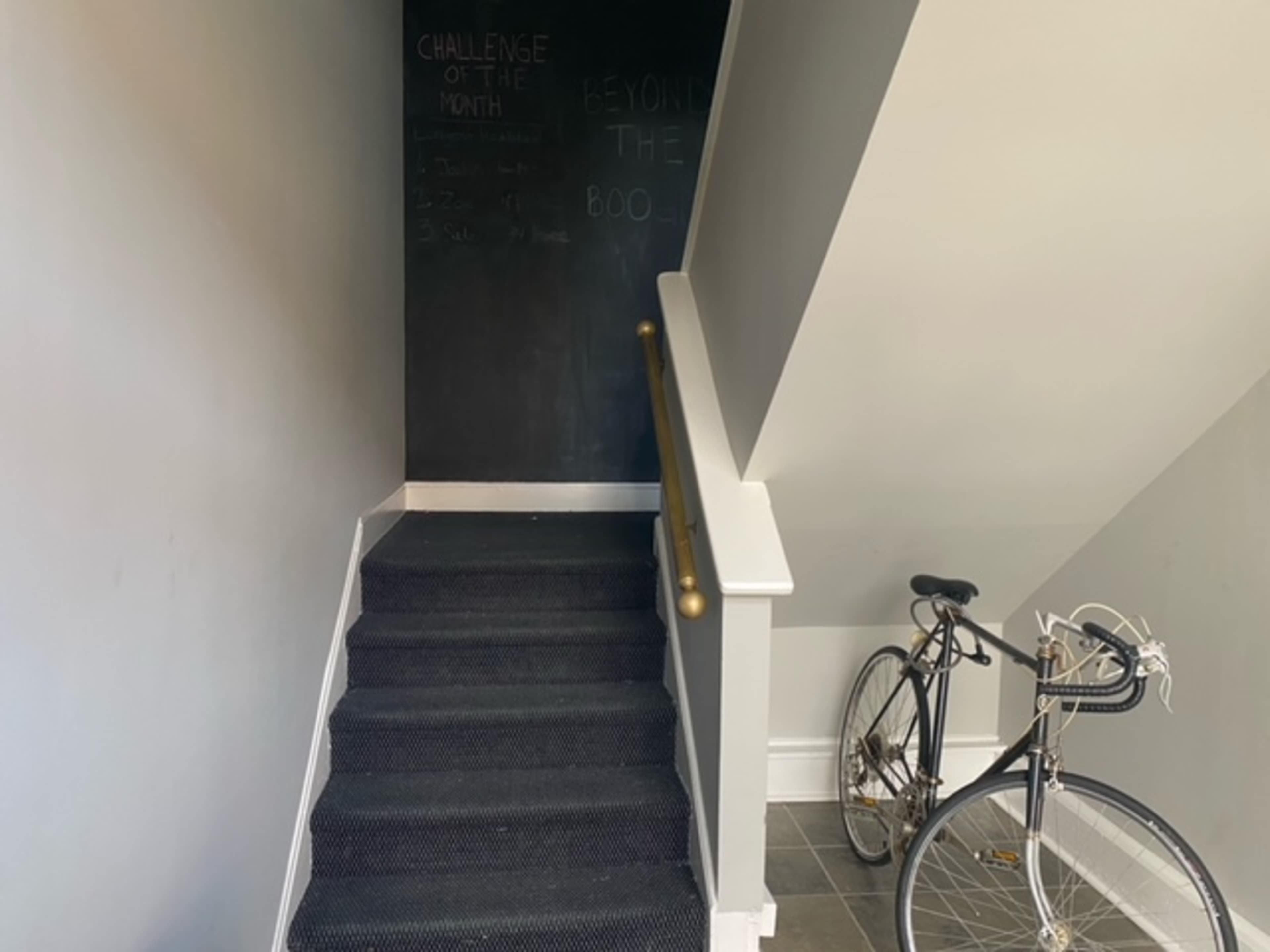A staircase leads up to a blackboard wall with writing, while a bicycle rests against the railing on the right.