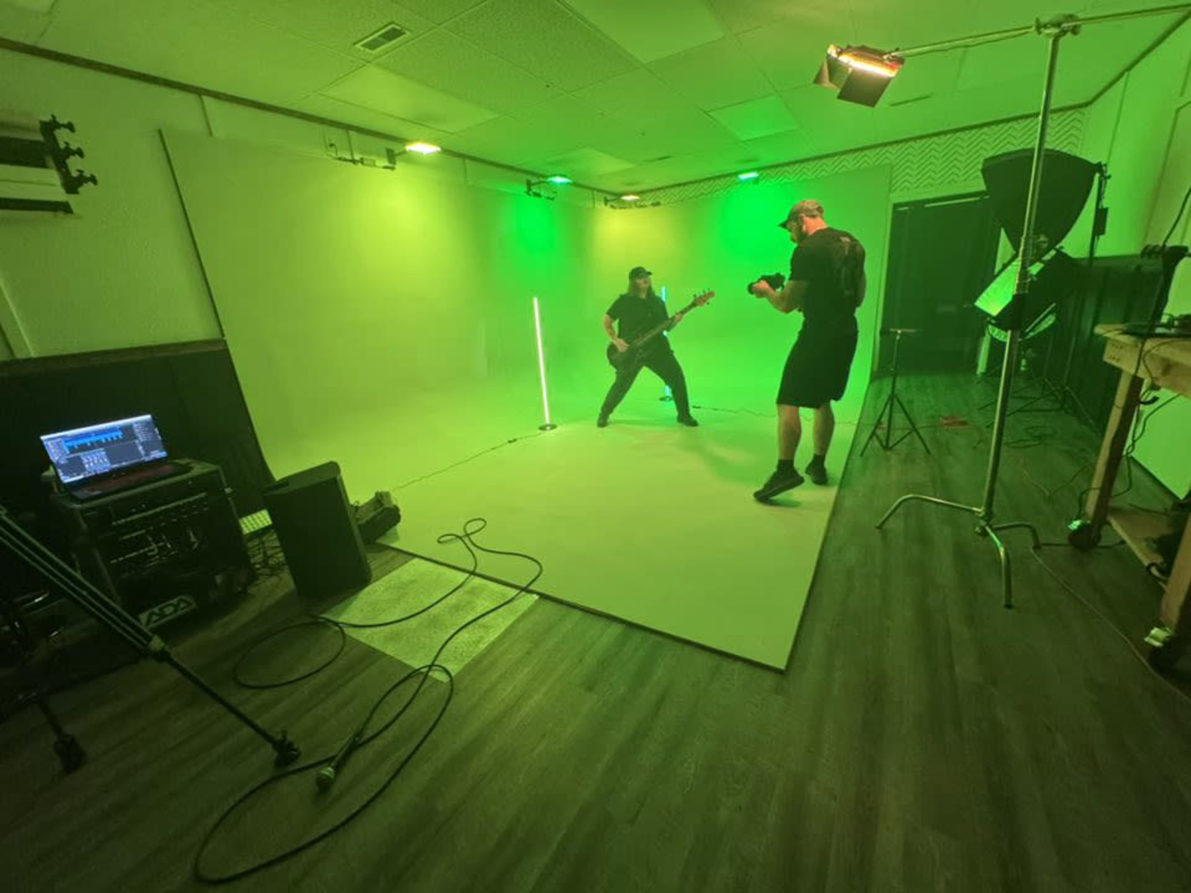 A musician plays guitar on a green-lit stage while another person records the performance with a camera.