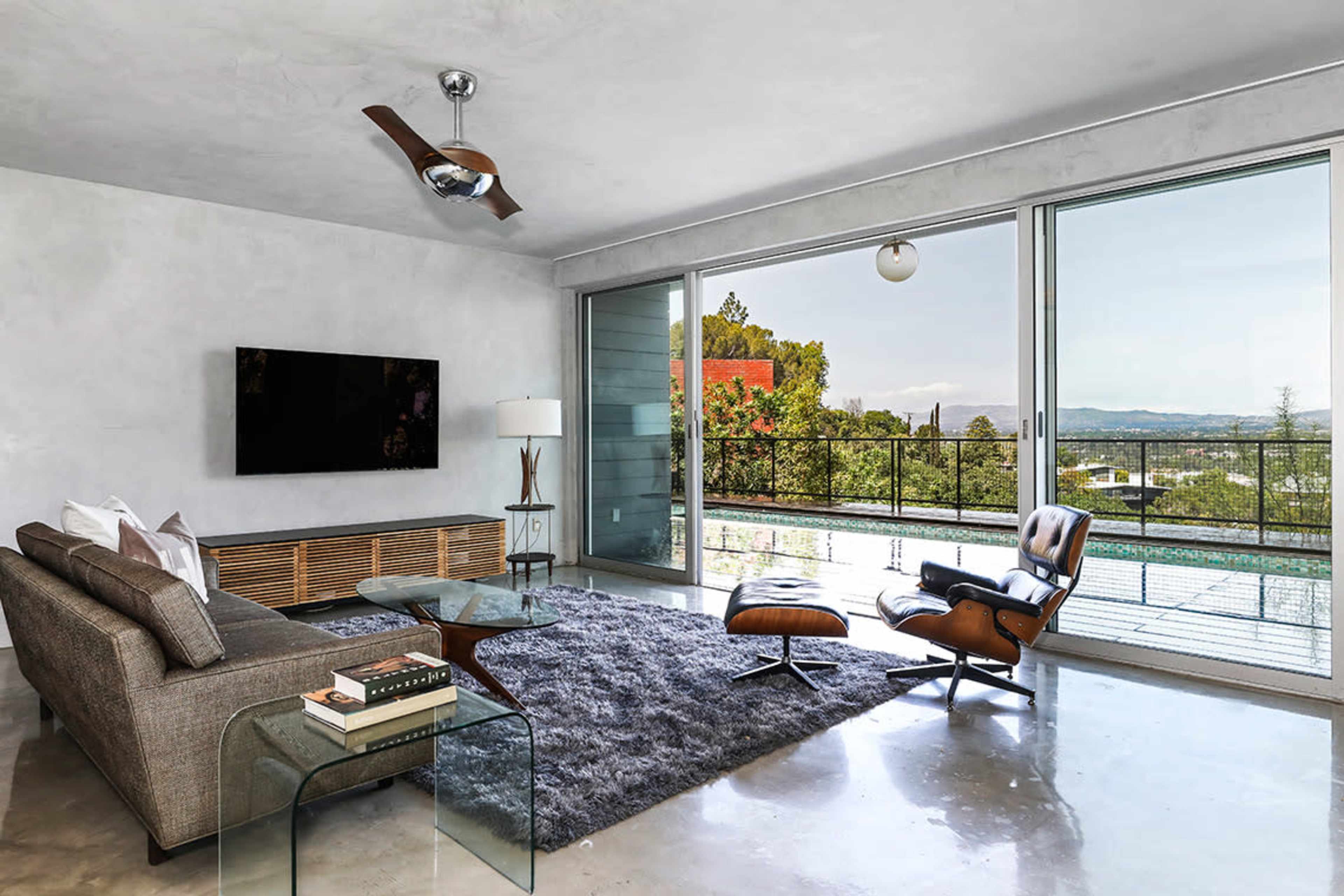 The living room features a modern design with a large window overlooking a scenic view, a sofa, an accent chair, a coffee table, and a flat-screen TV mounted on the wall.