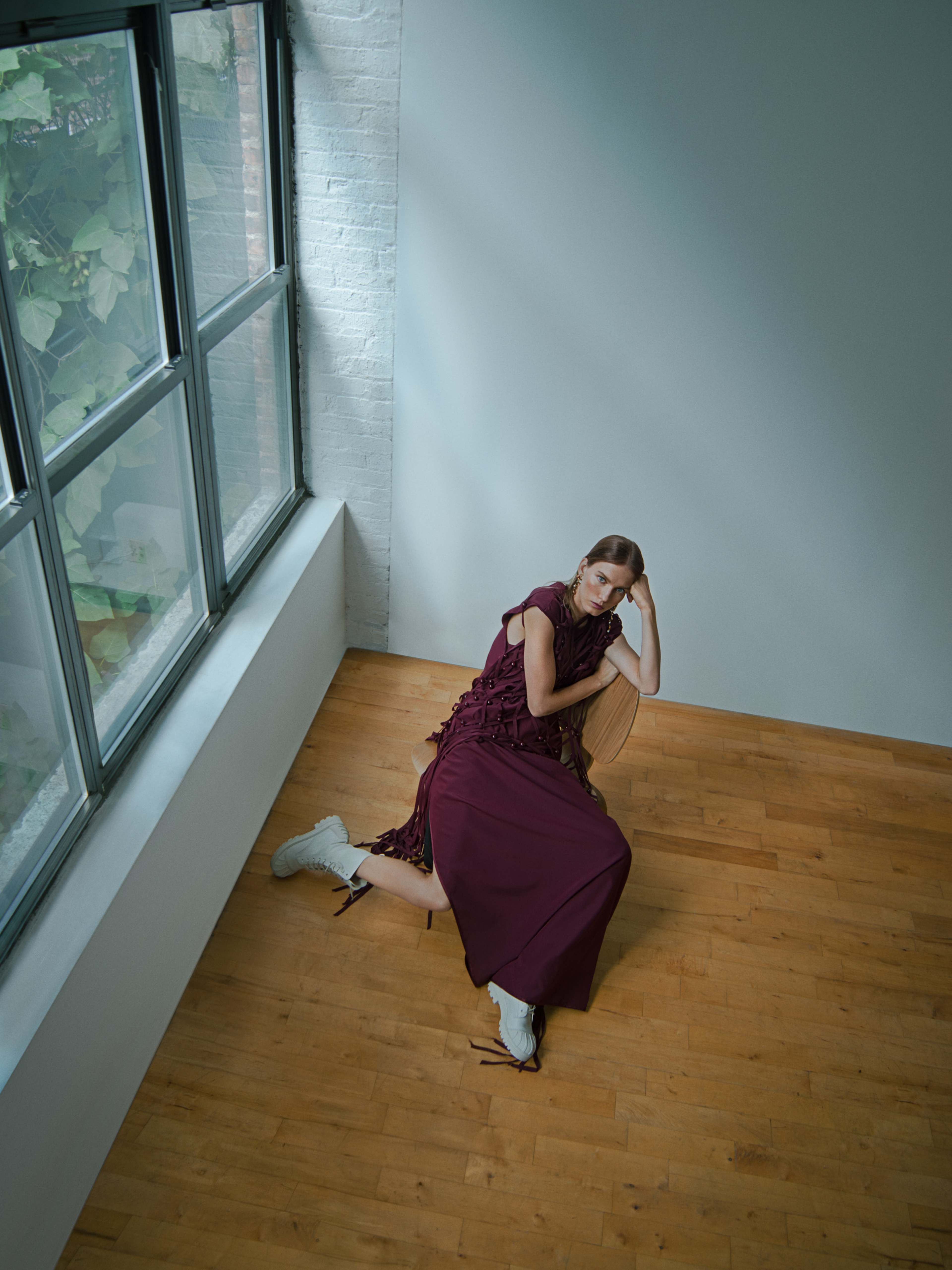 A model in a maroon dress sits on a wooden floor, leaning against a wall with large windows allowing natural light to illuminate the space.