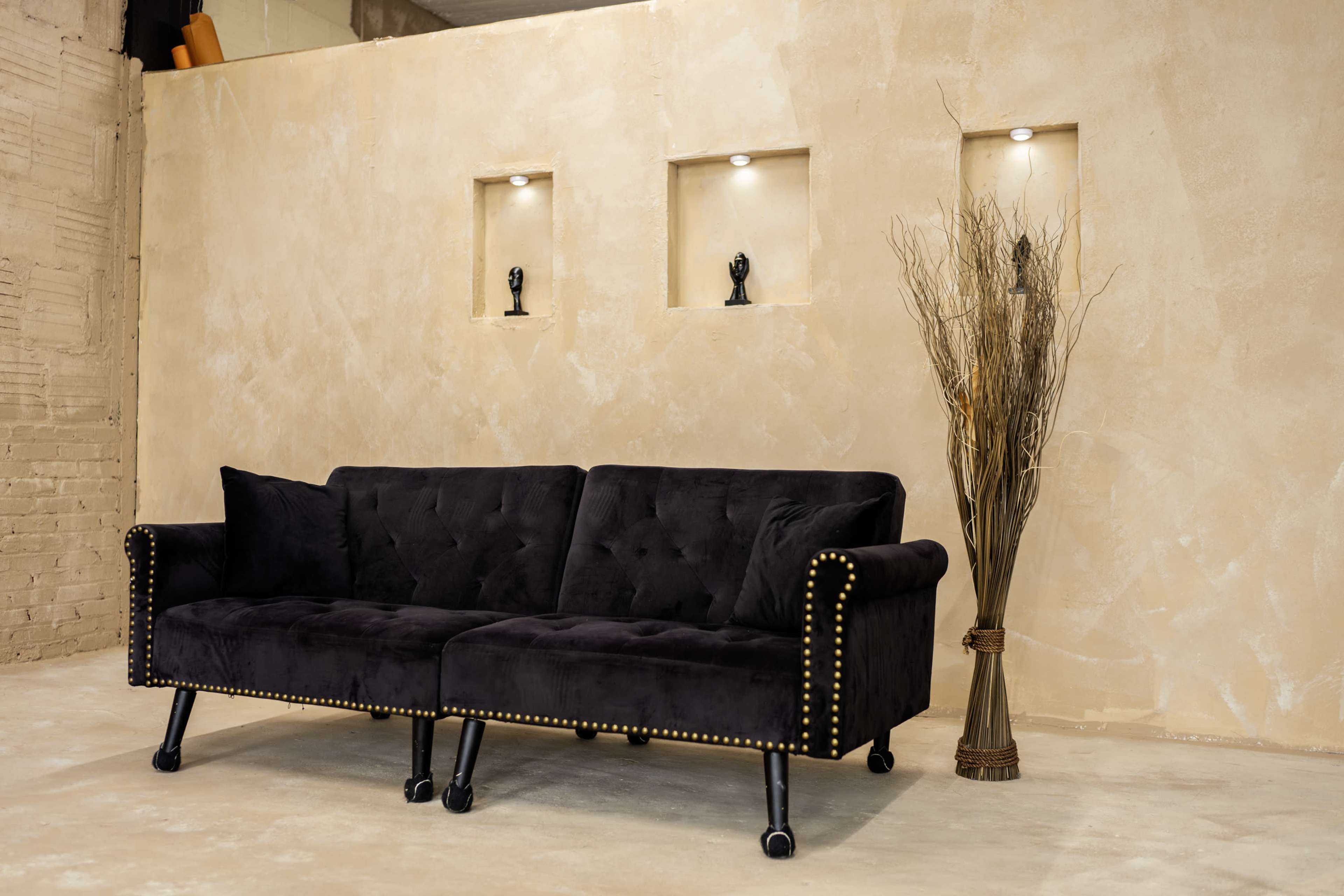The image shows a black velvet sofa with decorative nailhead trim, positioned against a light-colored textured wall that features three lit niches with small sculptures.