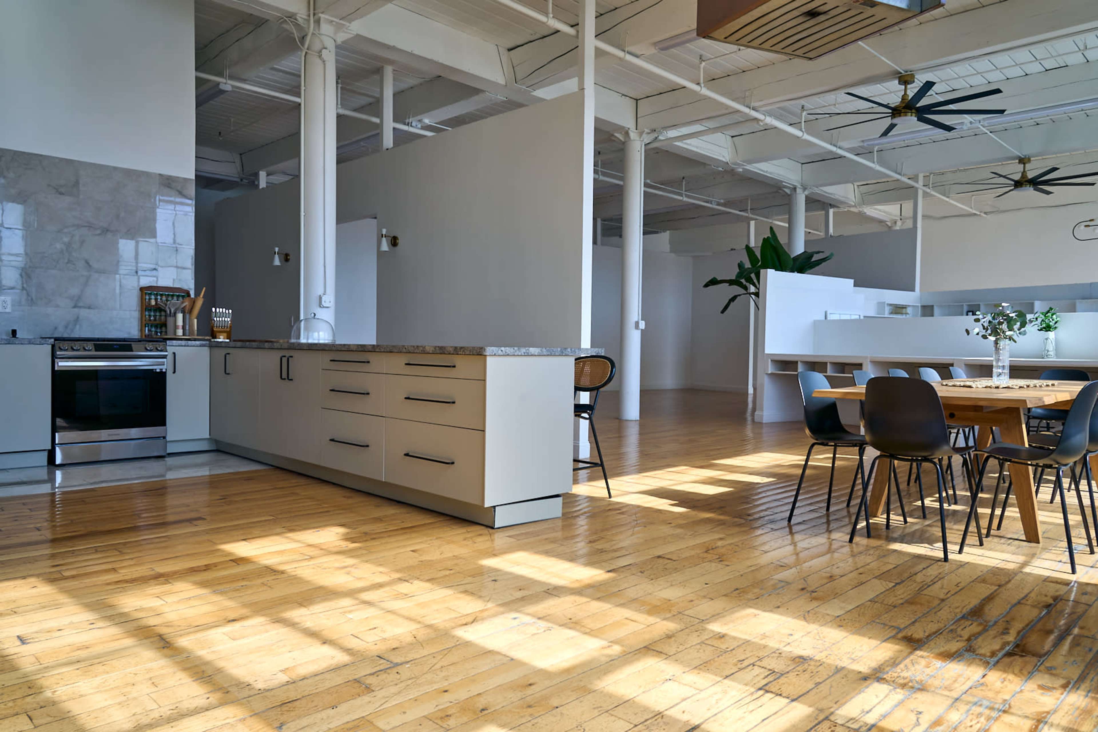 The image shows a spacious, modern kitchen and dining area with wooden floors, featuring an island counter, dining table, and large windows allowing natural light to fill the space.