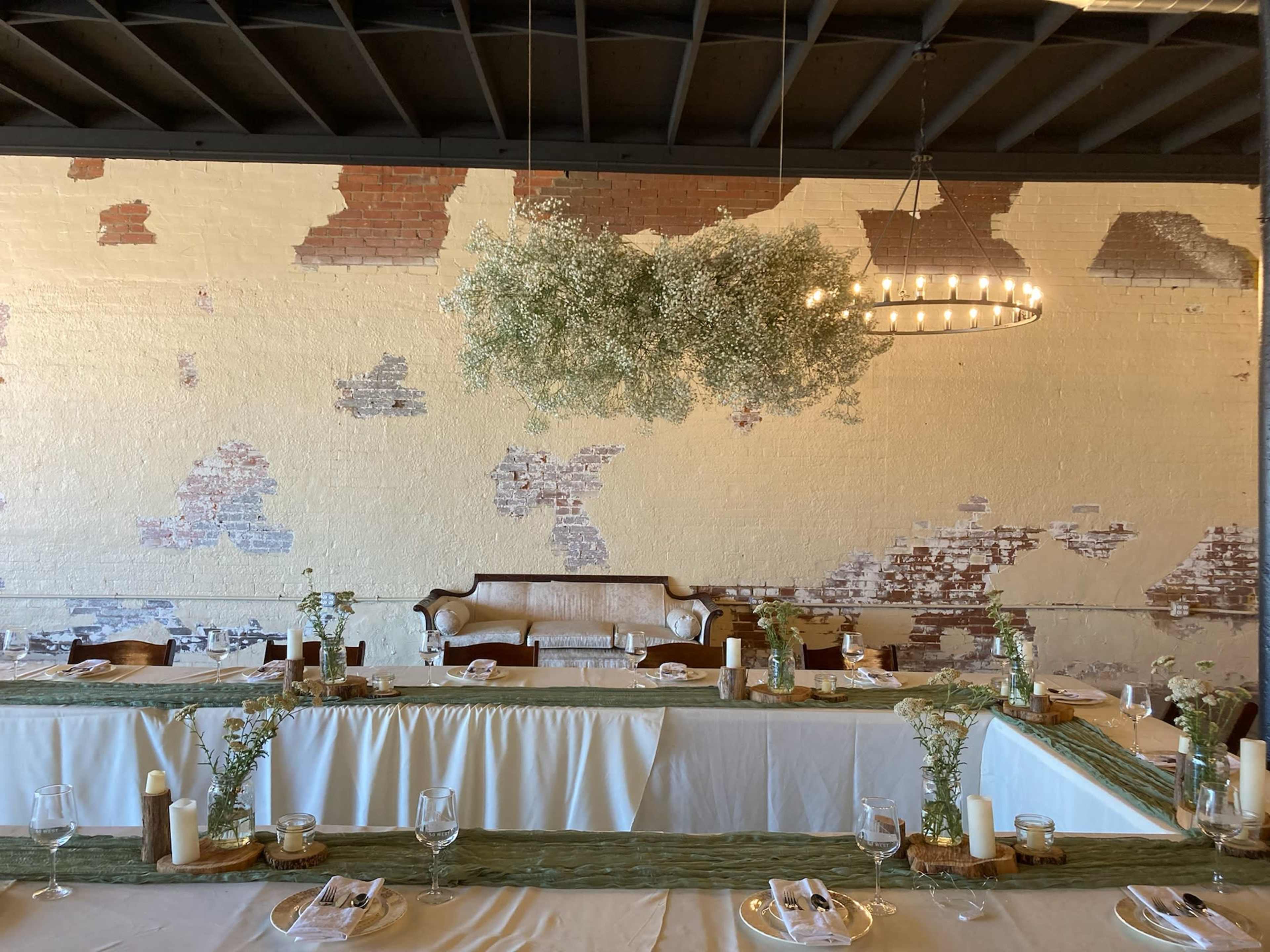A long, elegantly set dining table is arranged under a large, floral installation in a rustic venue with exposed brick walls.