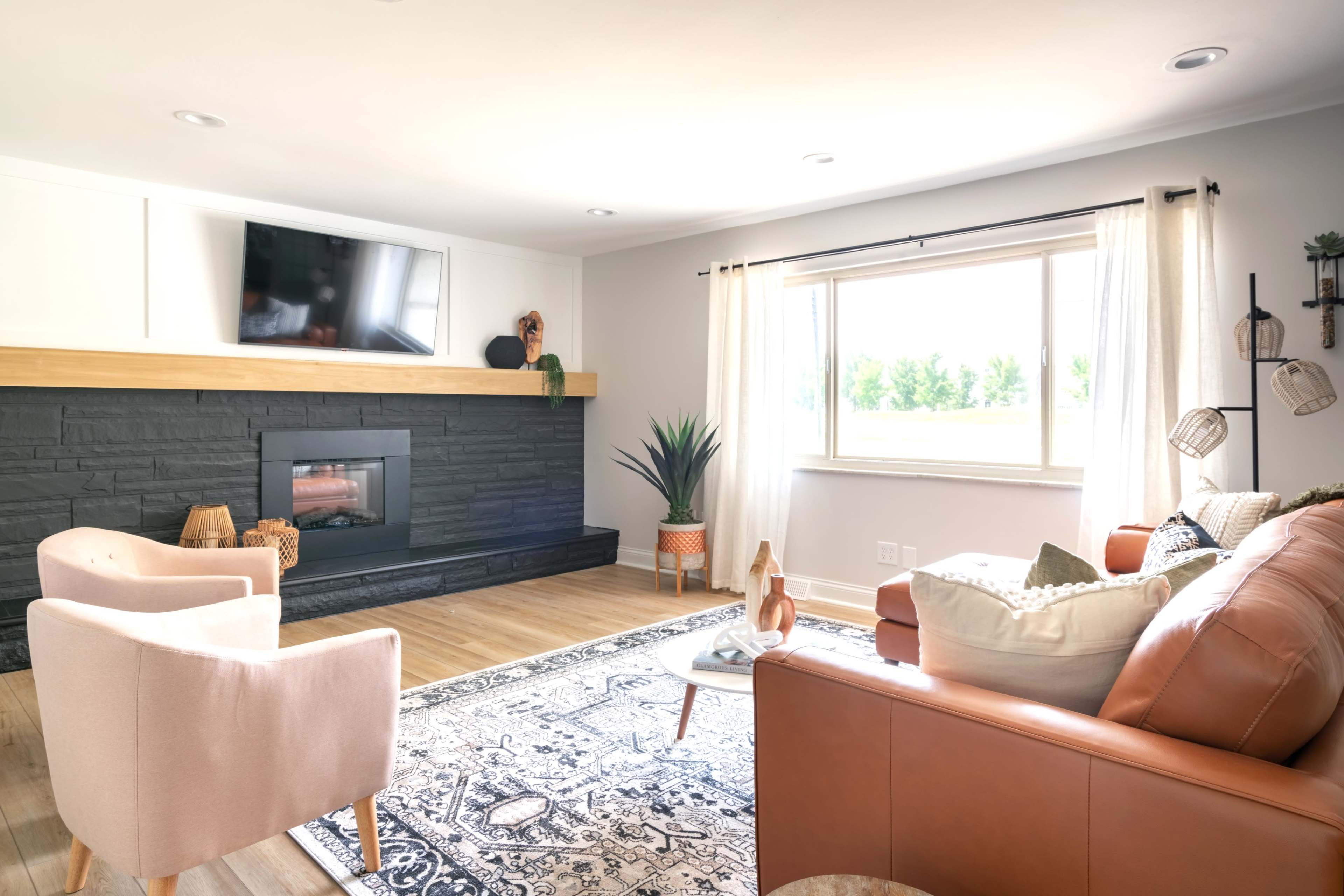 A modern living room features a television mounted above a black stone fireplace, light-colored furniture, and large windows letting in natural light.