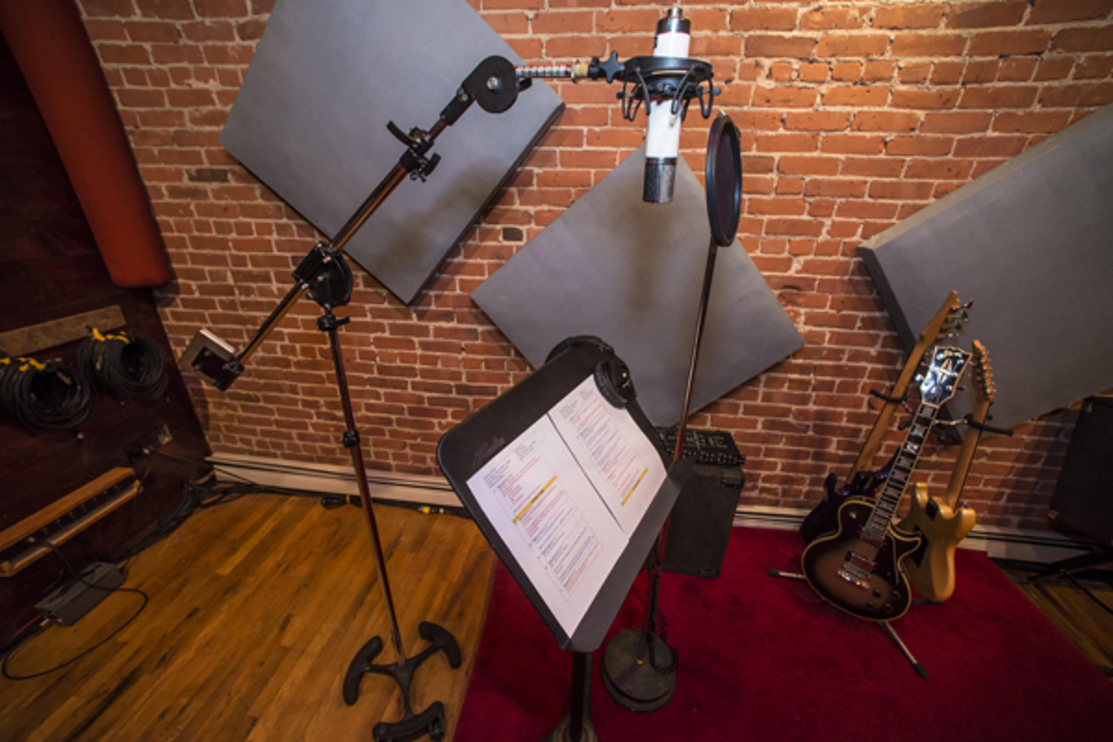 A recording studio setup featuring a microphone on a stand, a music stand with sheets, and an electric guitar resting against a wall with exposed brick.