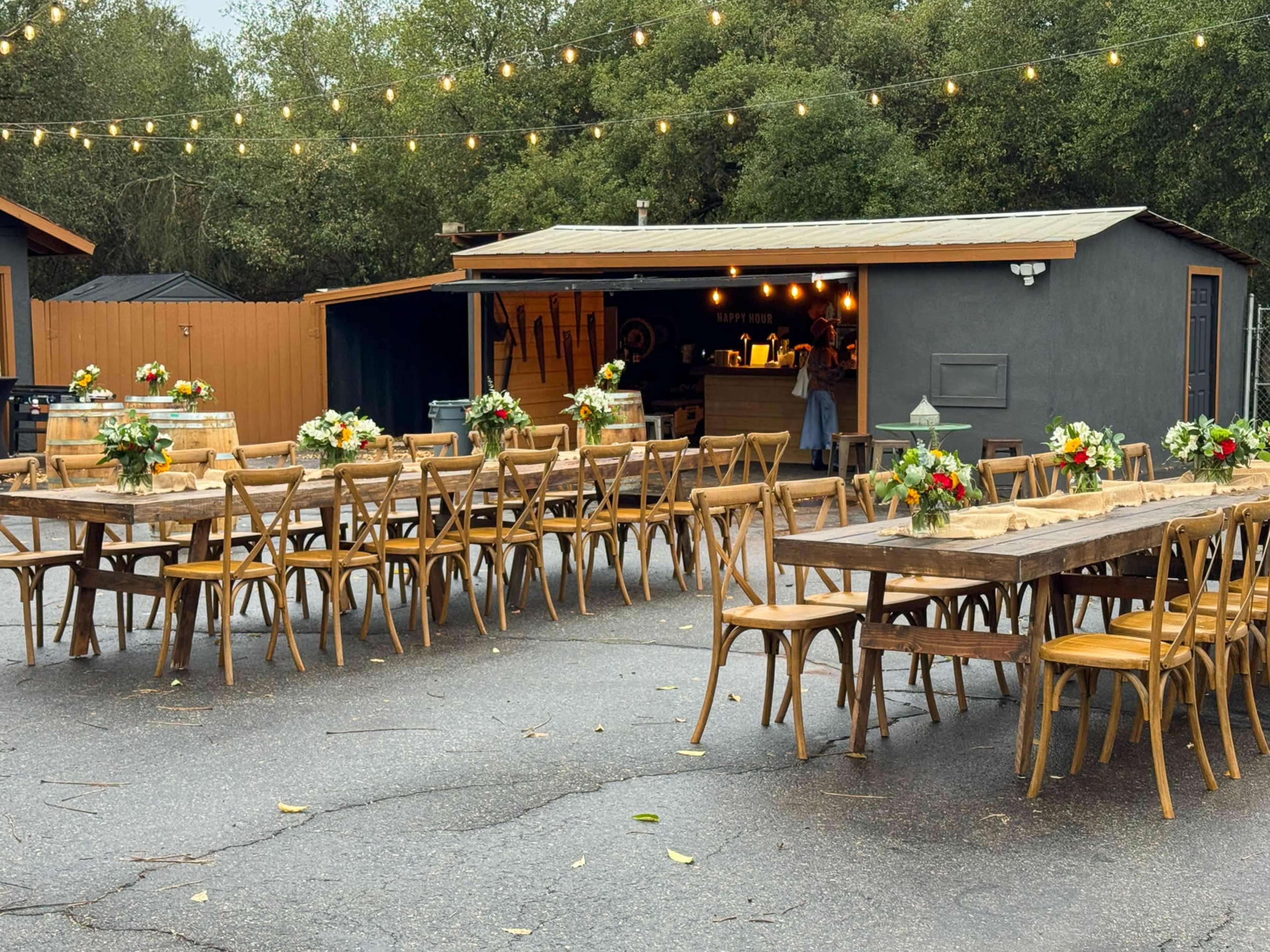 A spacious outdoor dining area features long wooden tables adorned with floral centerpieces, illuminated by string lights, adjacent to a small service building.