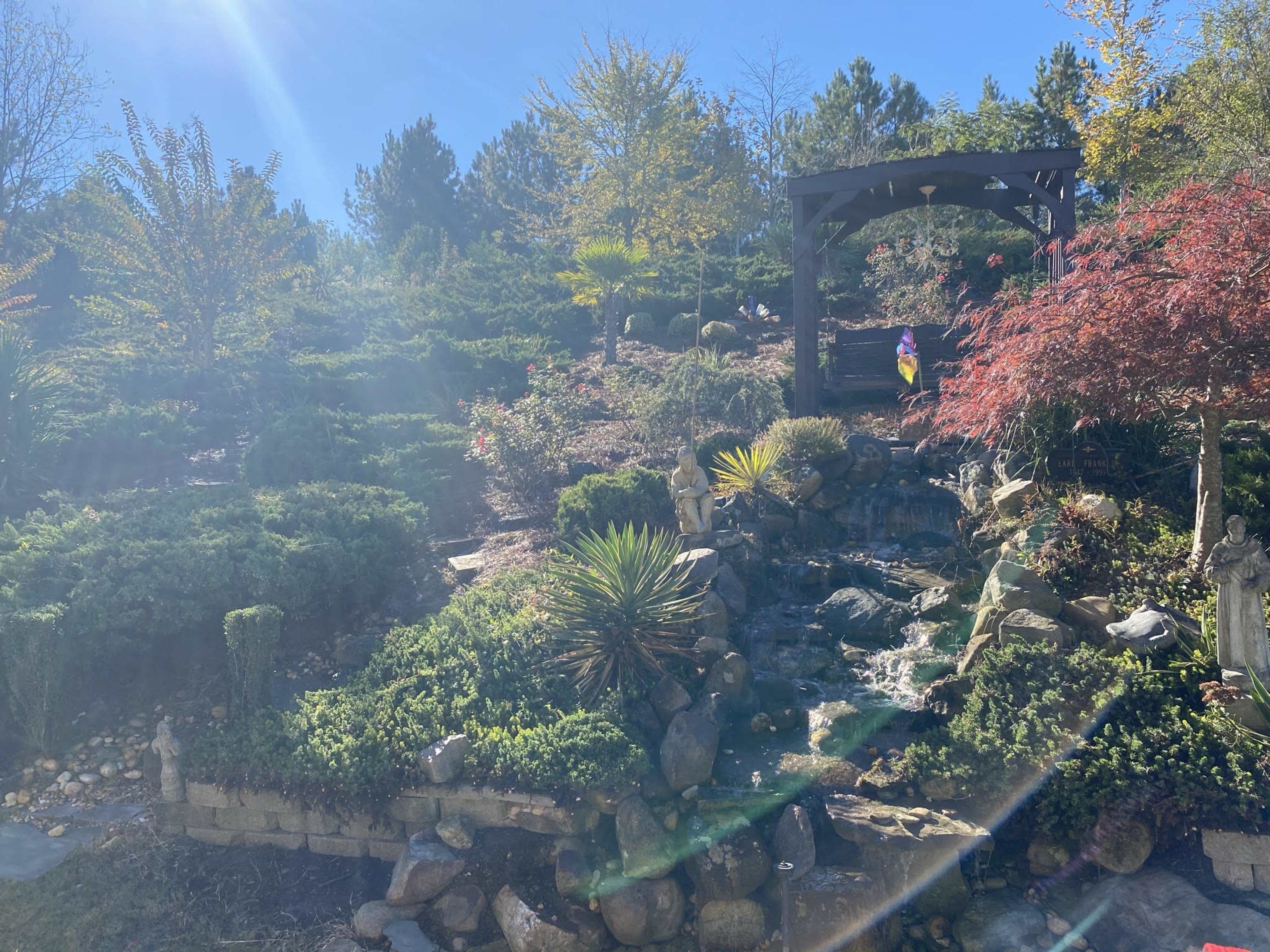 A landscaped garden with a stone waterfall, lush greenery, and an arbor in the background.