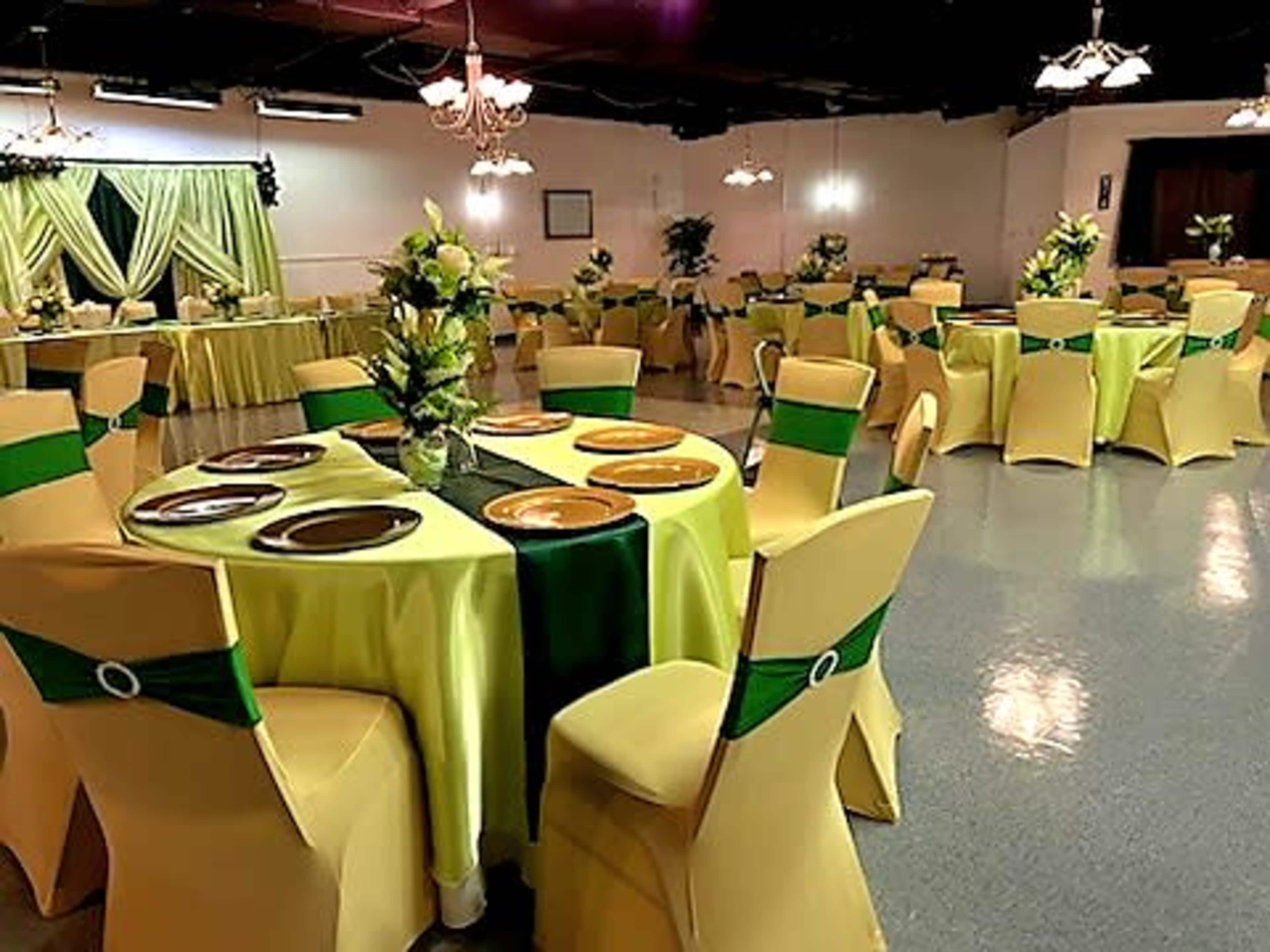 The image shows a banquet hall set with round tables draped in yellow tablecloths, featuring green sashes on the chairs, and decorated with floral centerpieces.