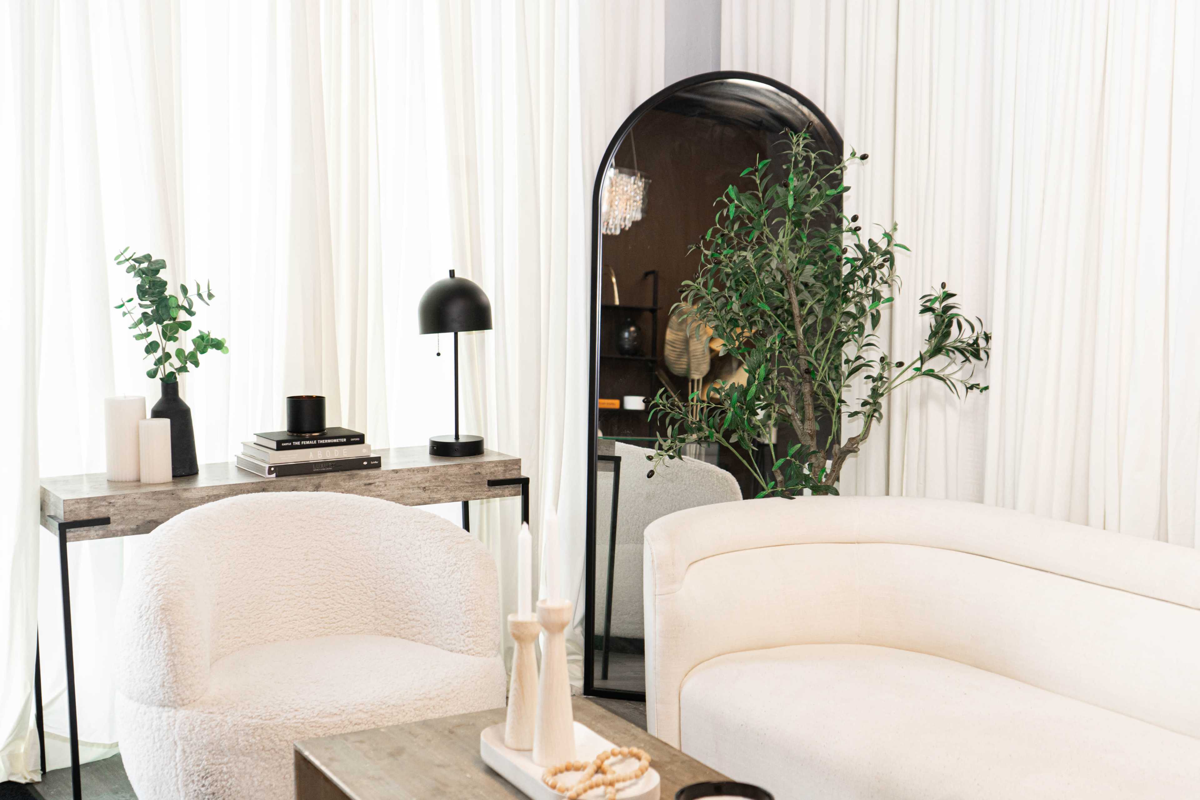 A modern living space featuring a curved white sofa, a small side table with a lamp, and a large mirror reflecting a plant and decorative items.
