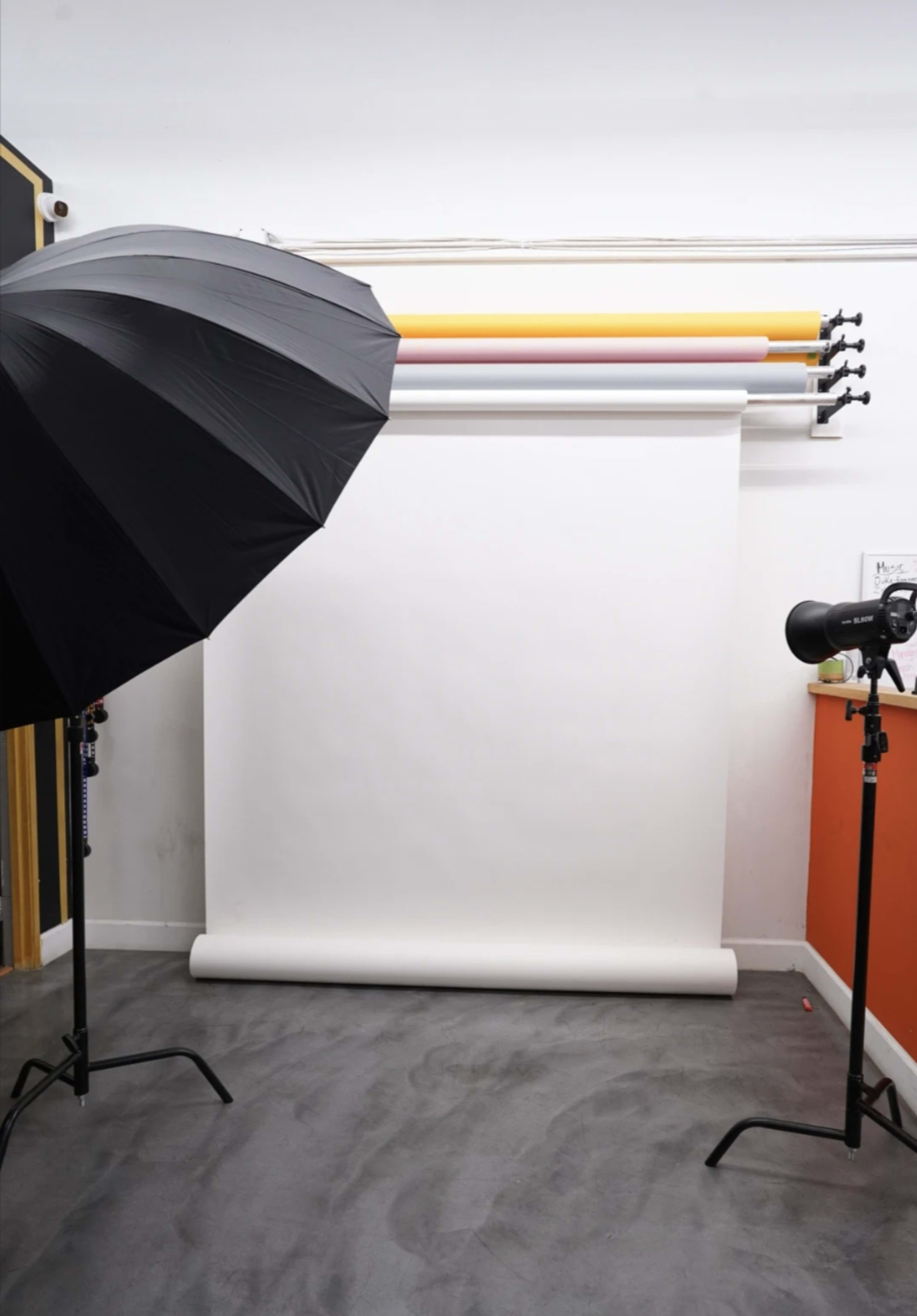 A photography studio features a white backdrop and two lighting setups, one with an umbrella and the other a spotlight.