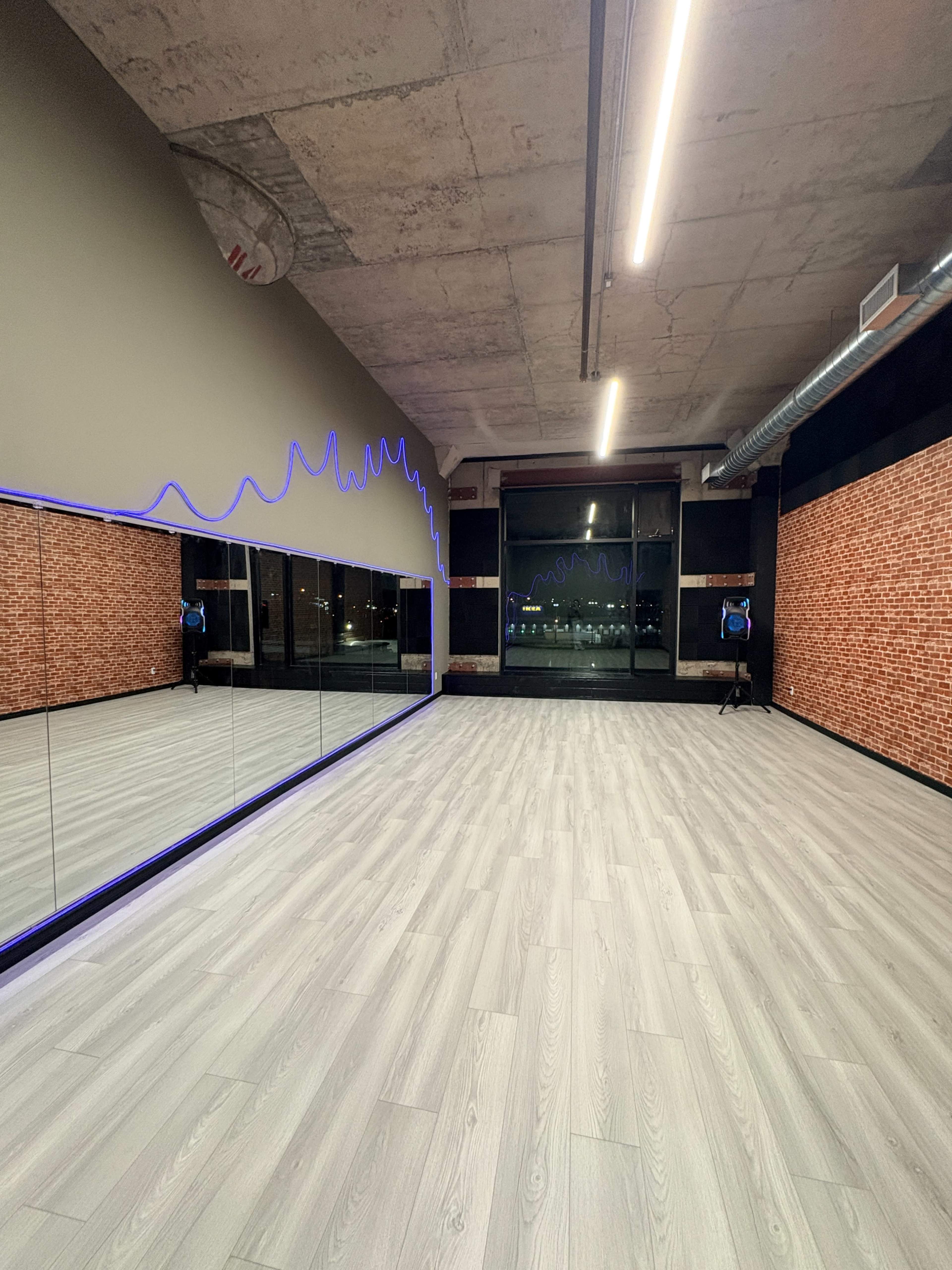 The image shows a spacious studio with a mirrored wall, modern flooring, and blue LED lighting along the walls, featuring large windows that provide natural light.
