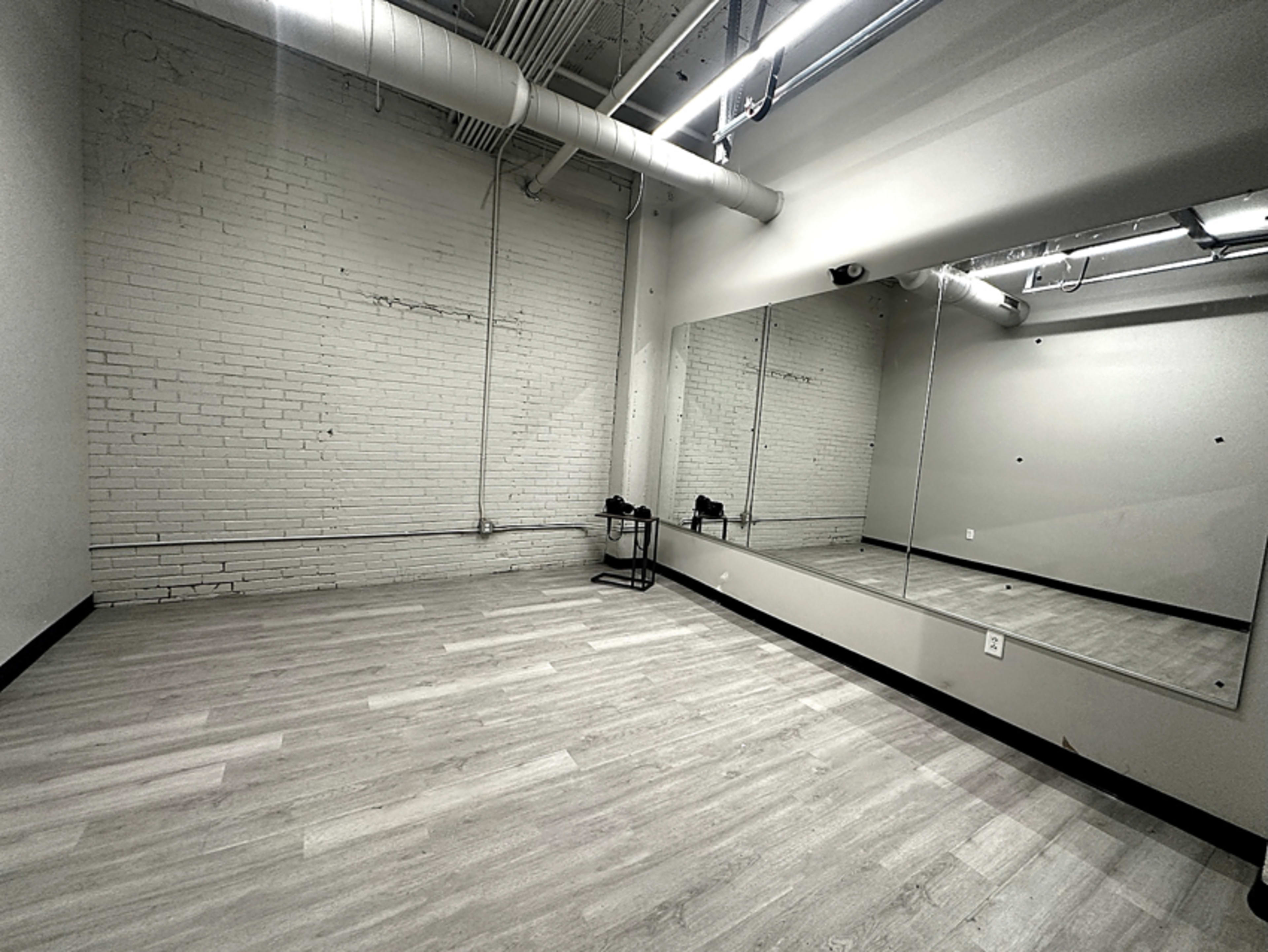 The image shows an empty room with light-colored wooden flooring, exposed brick walls, and a large mirror along one side.