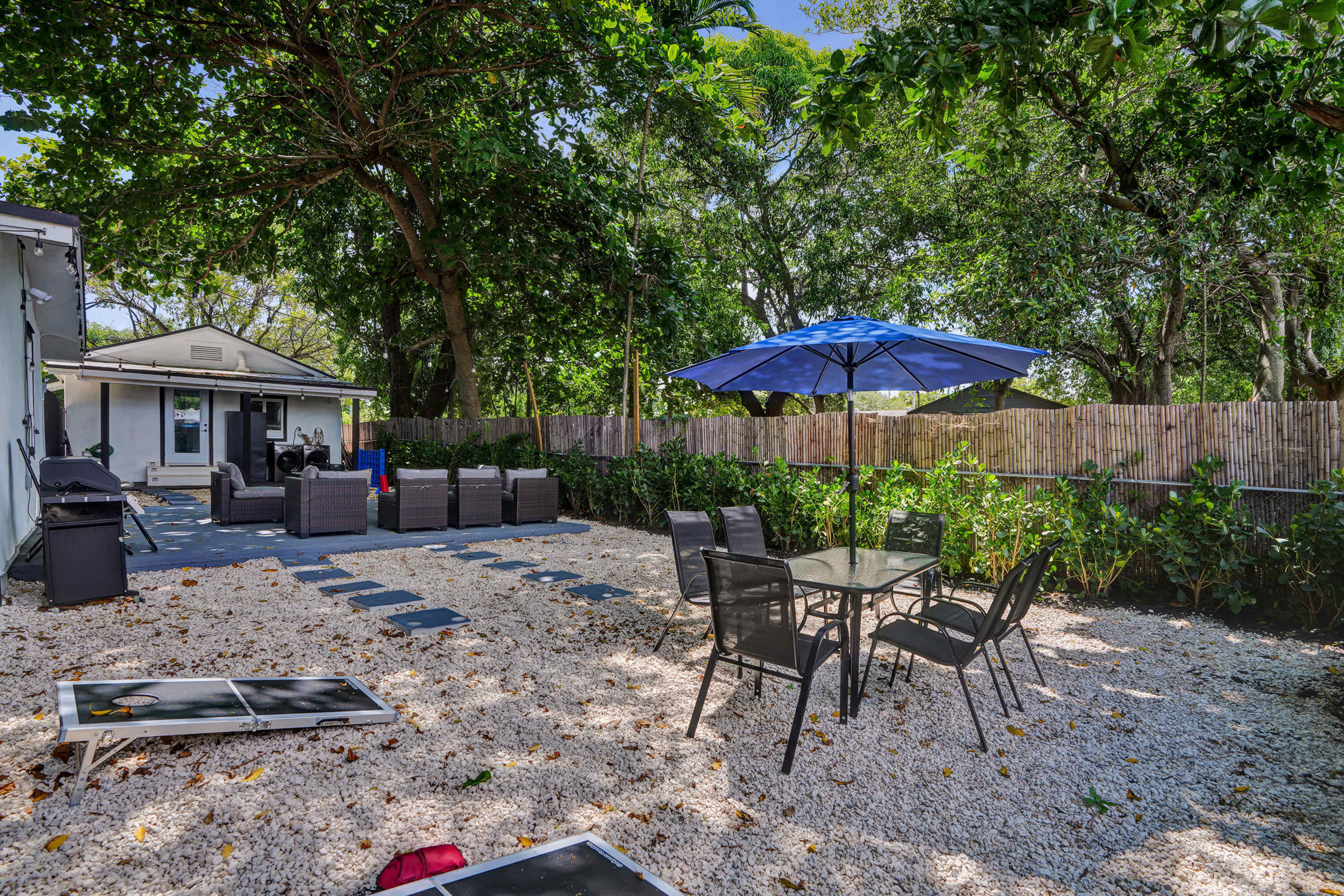 The image displays a landscaped outdoor area featuring a gravel surface, a glass-top dining table with chairs, a large blue umbrella, and a seating arrangement under tree cover.