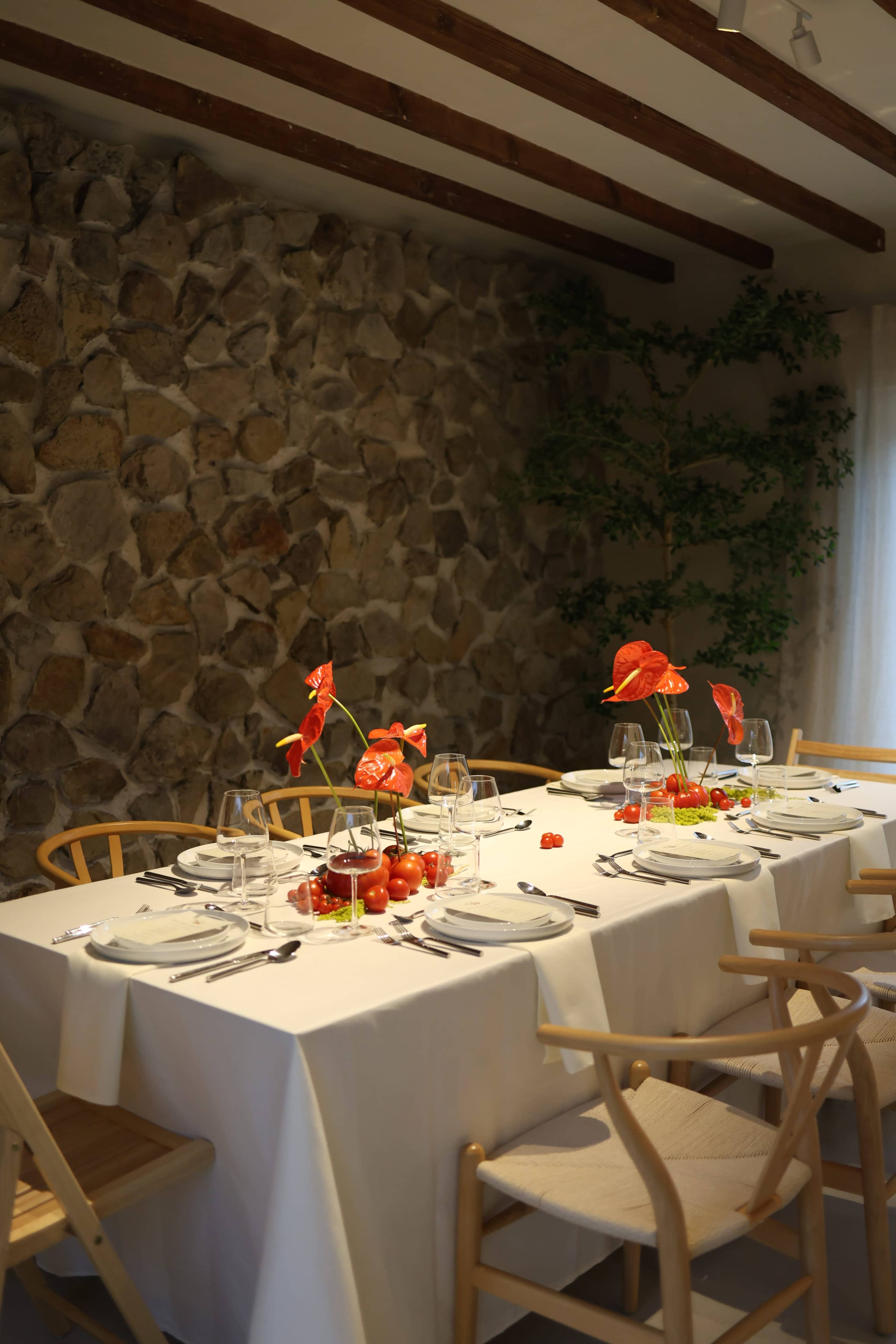 A dining table is elegantly set with white tablecloths, glassware, plates, and red floral centerpieces against a stone wall.