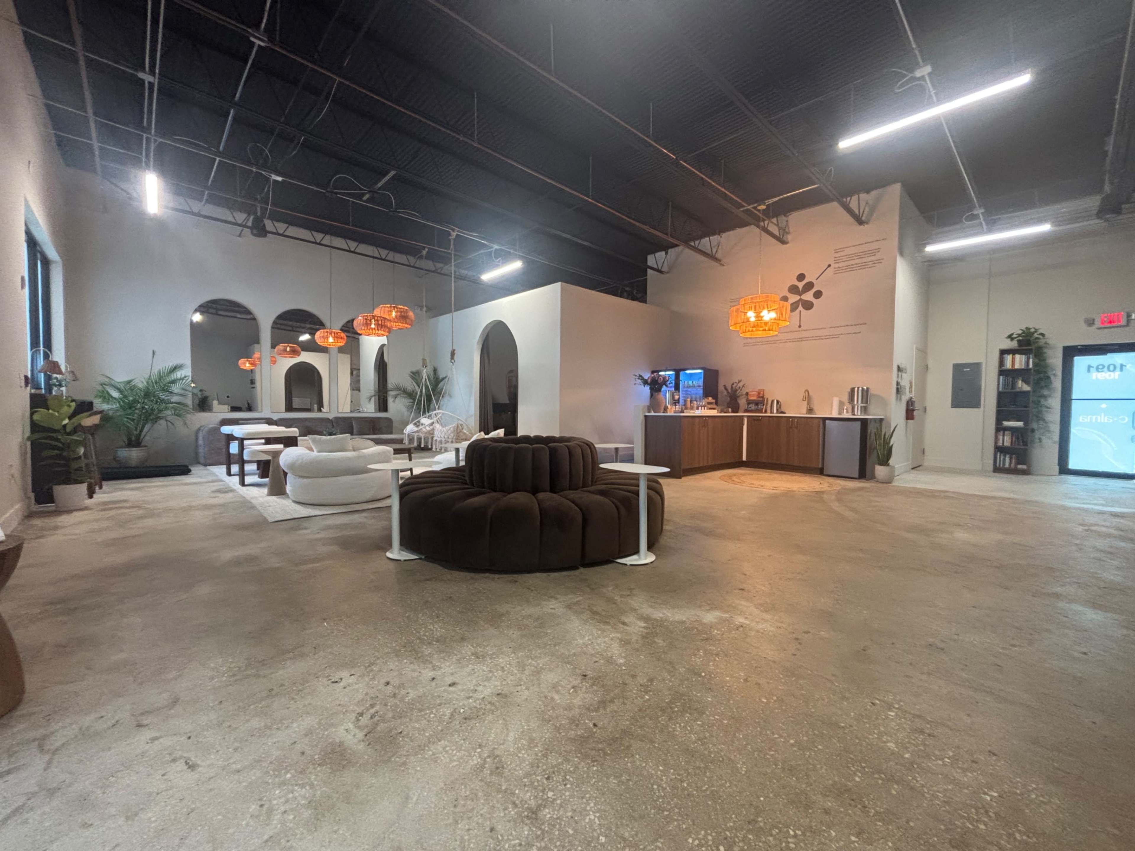 The image shows a spacious, modern interior with a welcoming lounge area featuring a circular brown sofa and several large pendant lights, along with a functional kitchen and dining space.