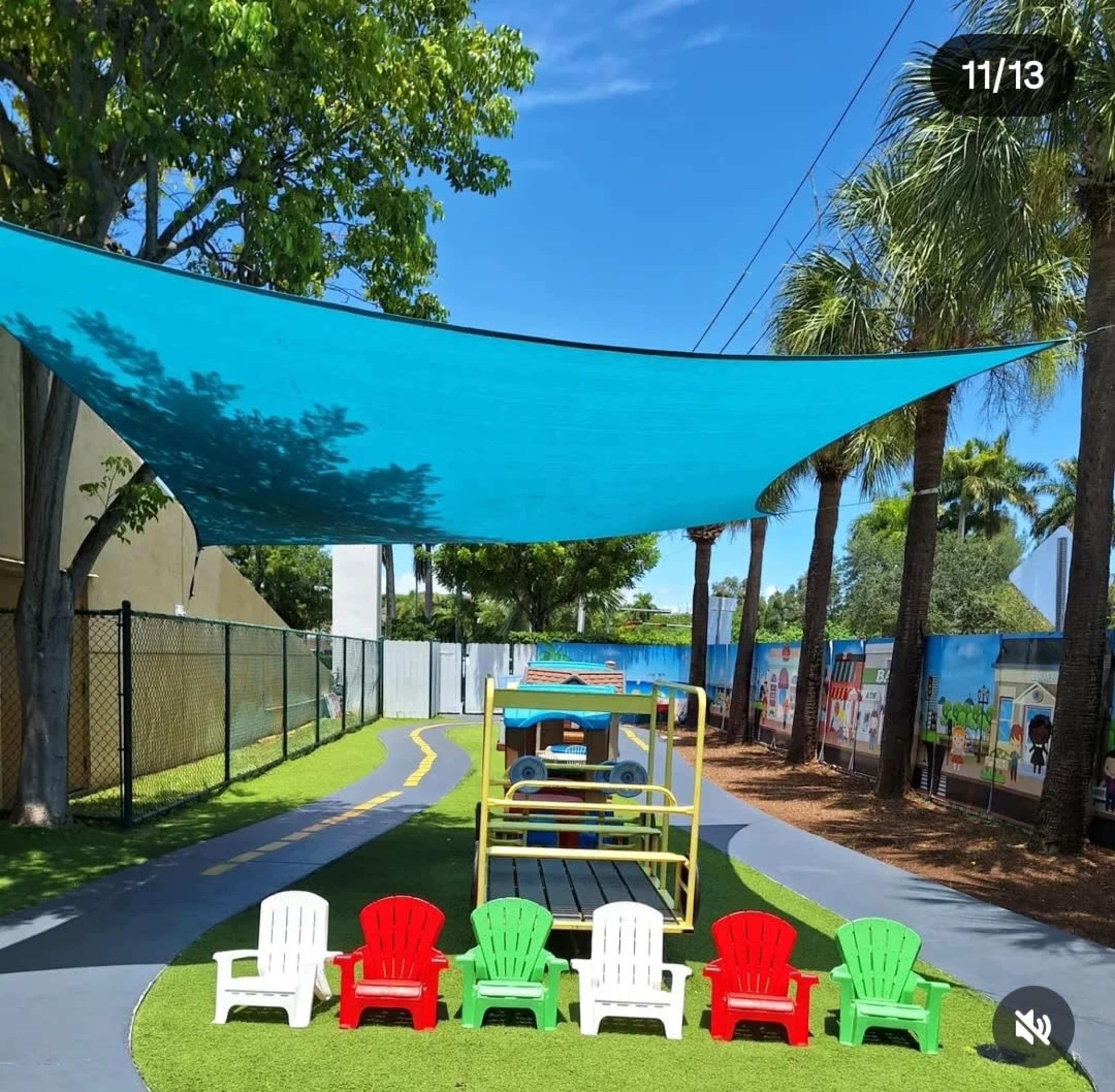 The image shows a playground area with colorful chairs, a blue shade sail overhead, and a play structure on a grassy surface surrounded by palm trees.