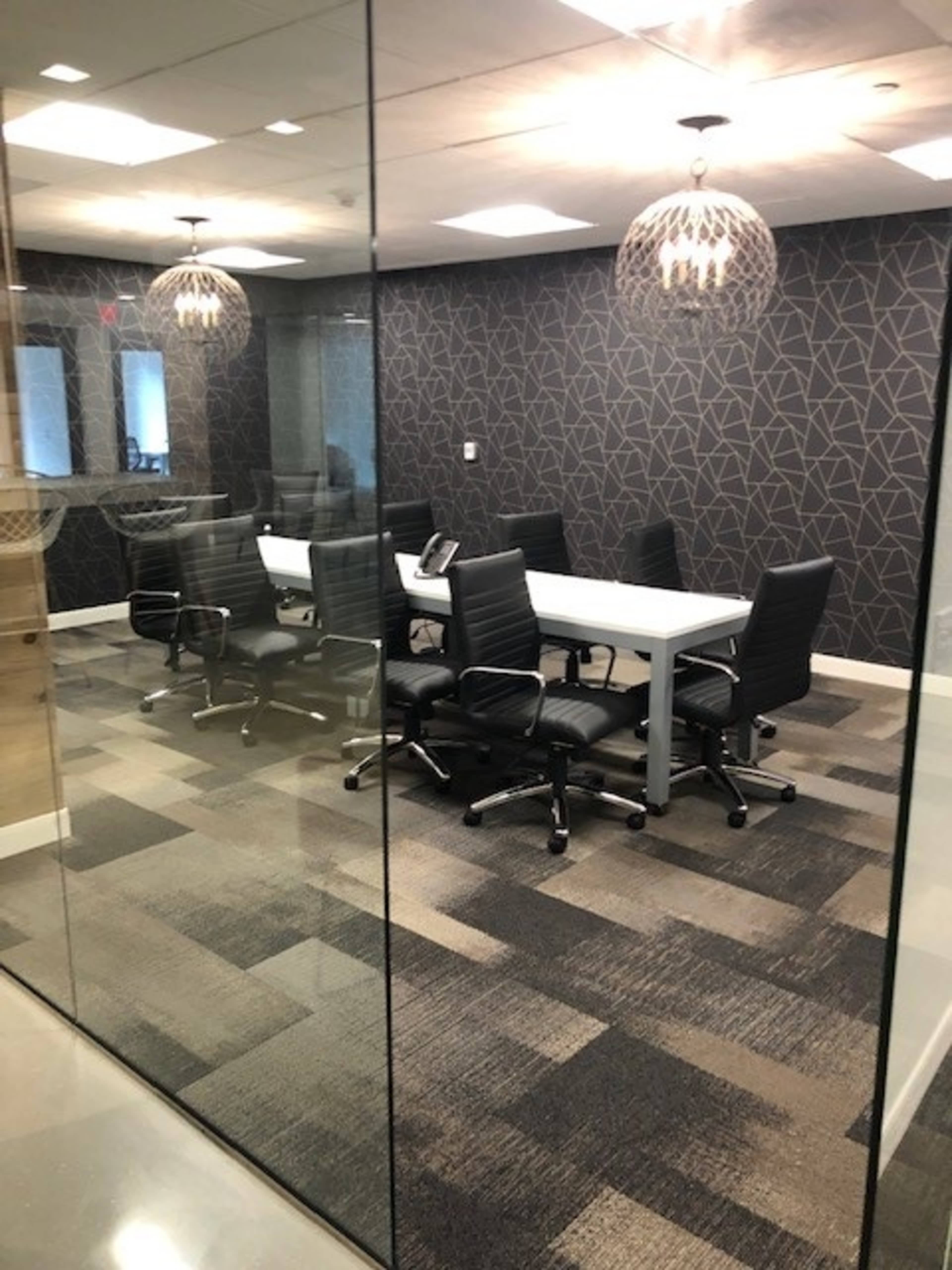 A glass-walled conference room features a large table surrounded by black office chairs and is illuminated by pendant lights.
