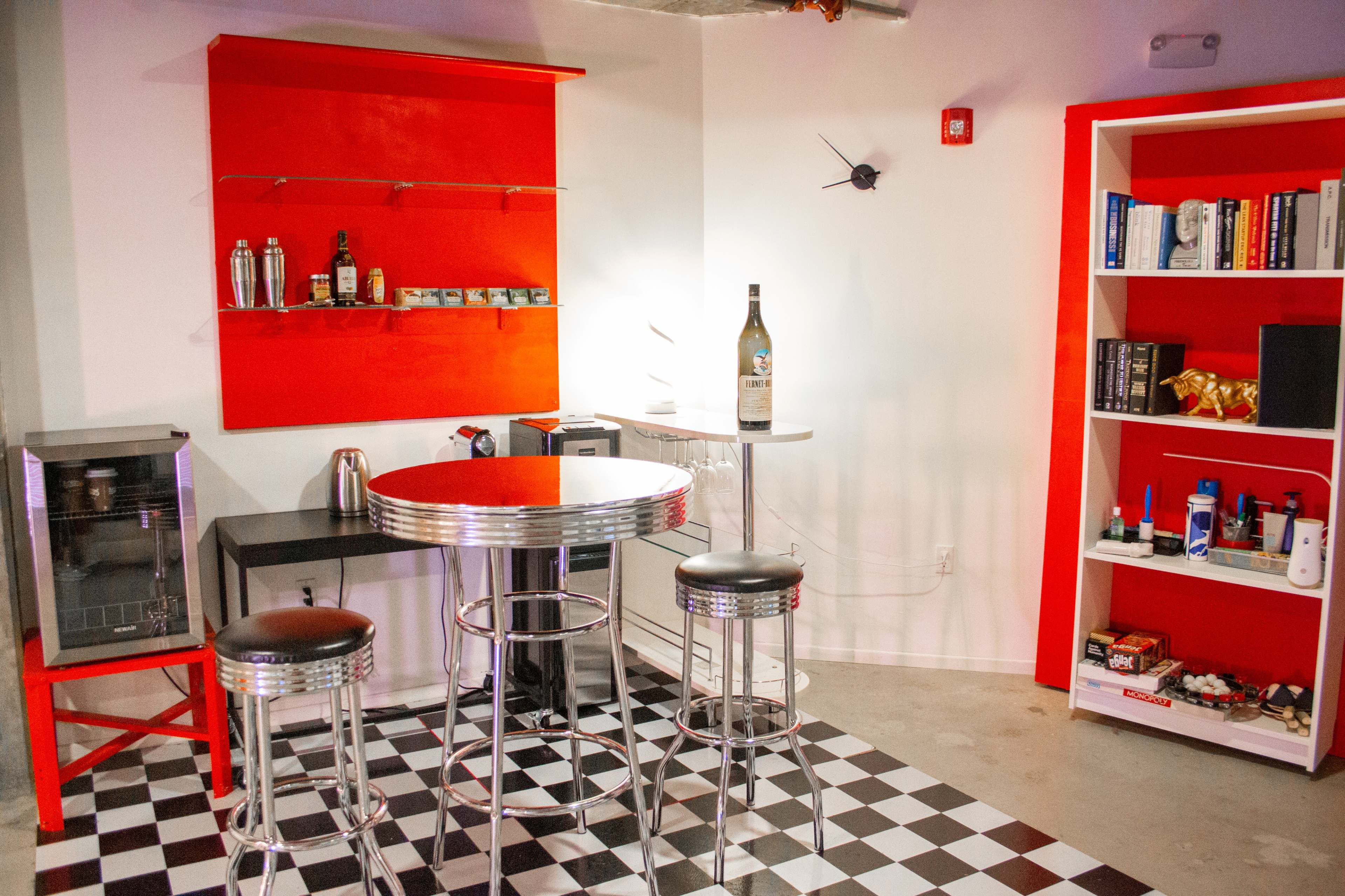 A modern bar area with a round chrome table, three bar stools, and a red shelf and bookshelf against white walls, with a checkerboard floor.