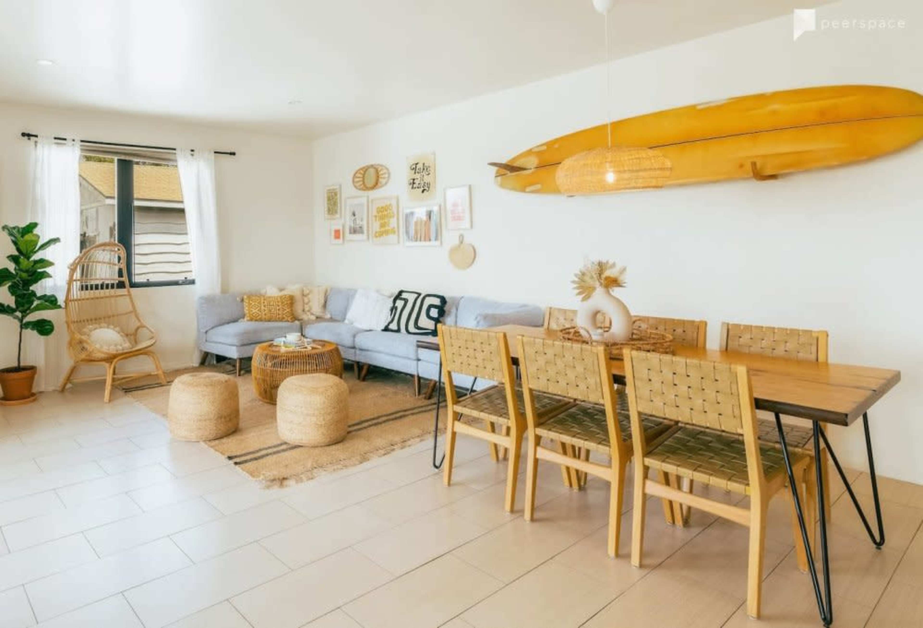 A bright living space with a blue sofa, wooden dining table with six chairs, and decorative surfboards hanging on the wall.