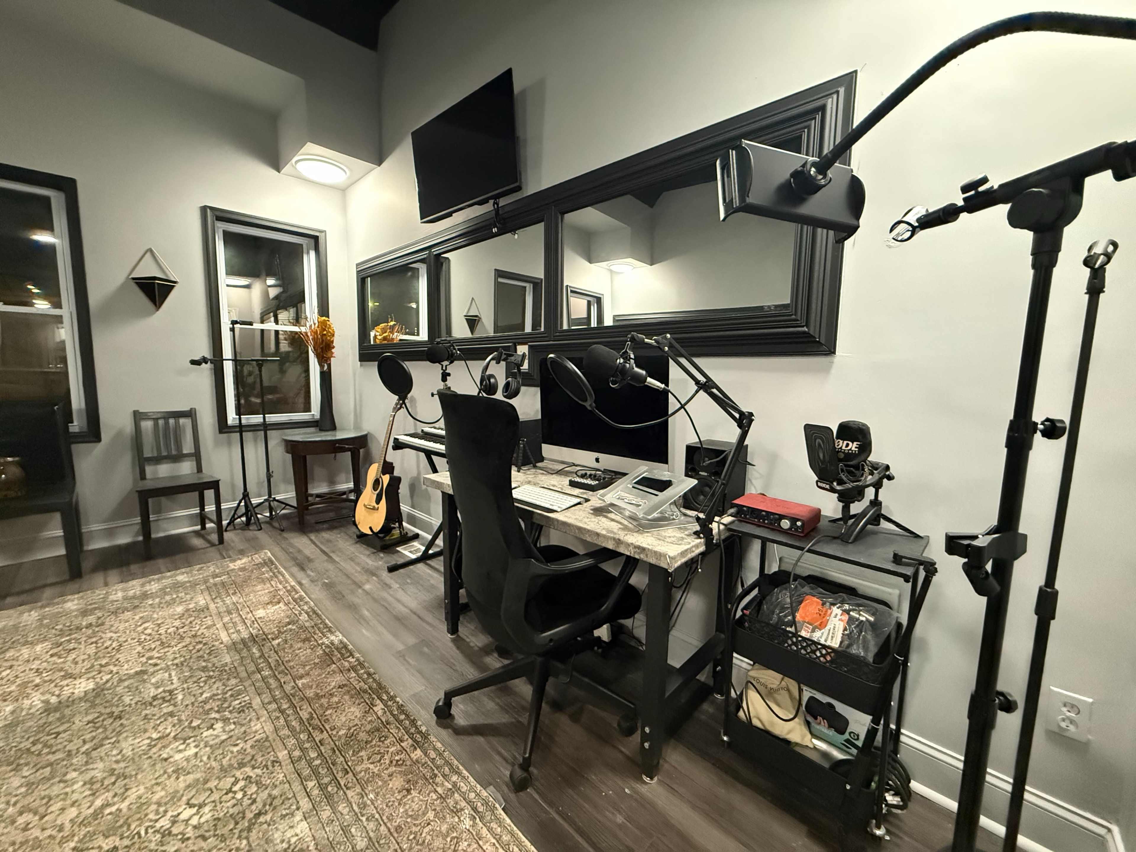 The image shows a modern recording or media studio featuring a desk with a computer, microphones, and a guitar, surrounded by acoustic panels and dim lighting.