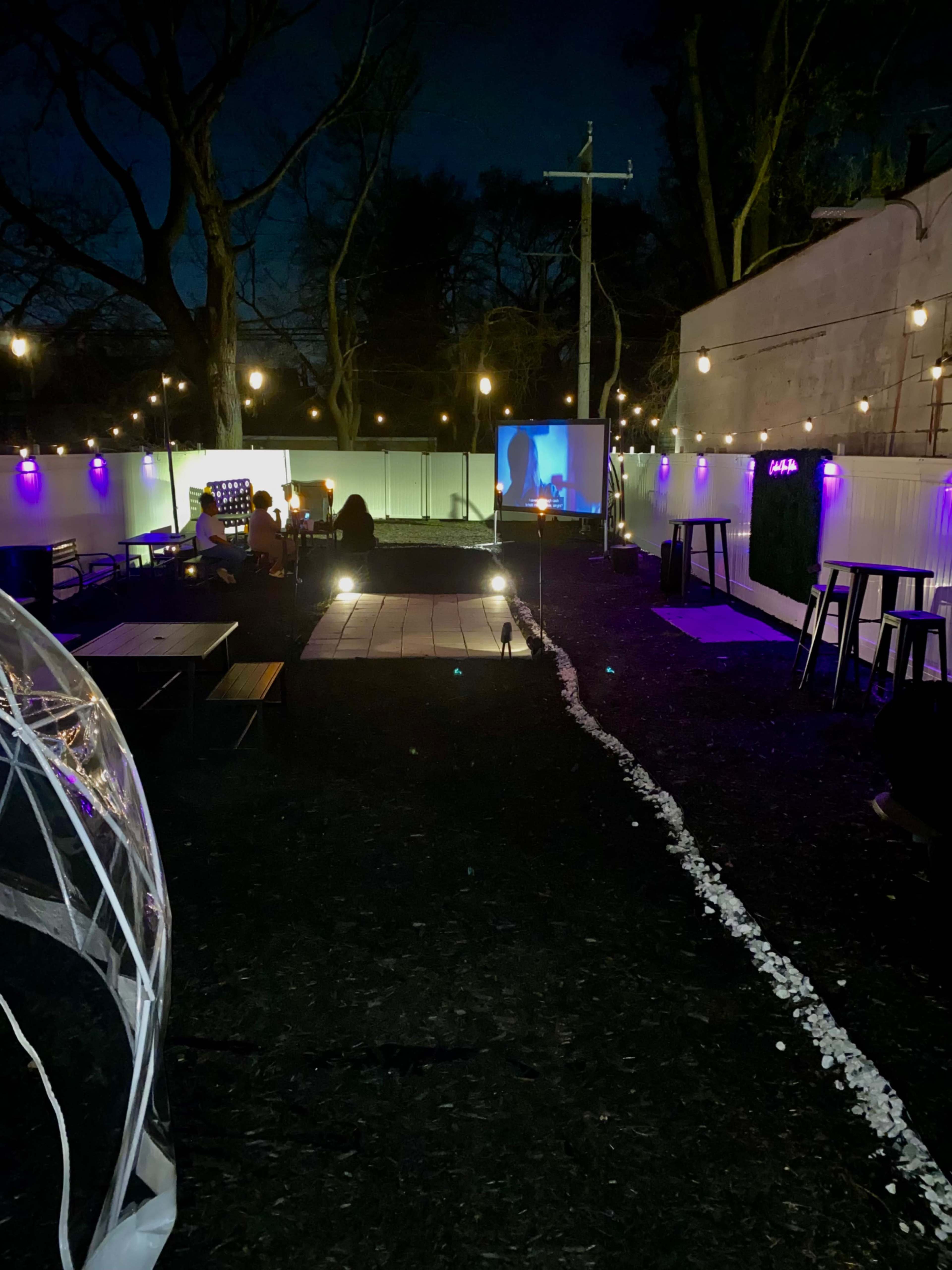 The image shows an outdoor space at night with a projection screen, seating areas, and string lights illuminating the pathway.