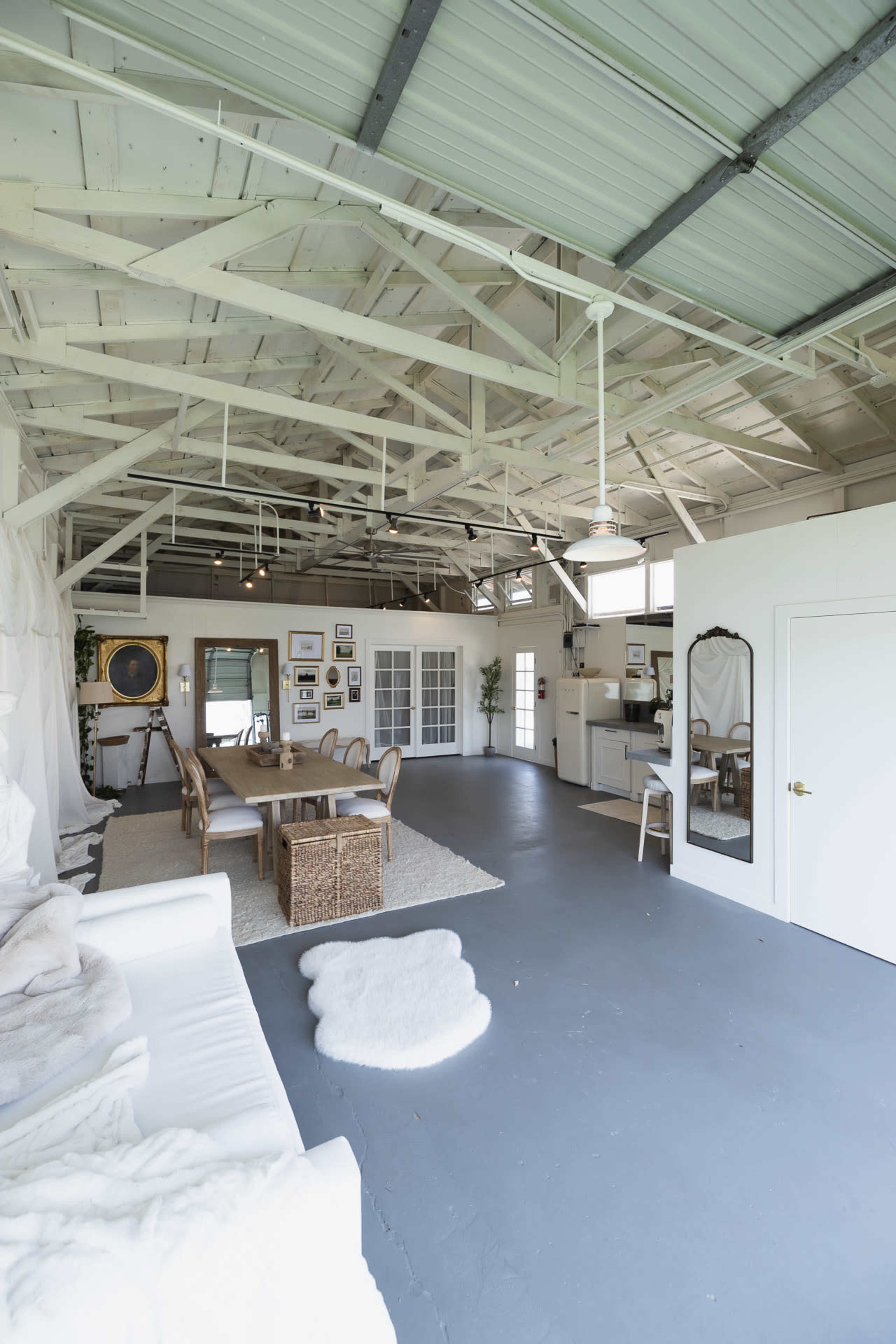 The image depicts a spacious, modern interior featuring a white sofa, a wooden coffee table, and various wall decorations under a high ceiling with exposed beams.
