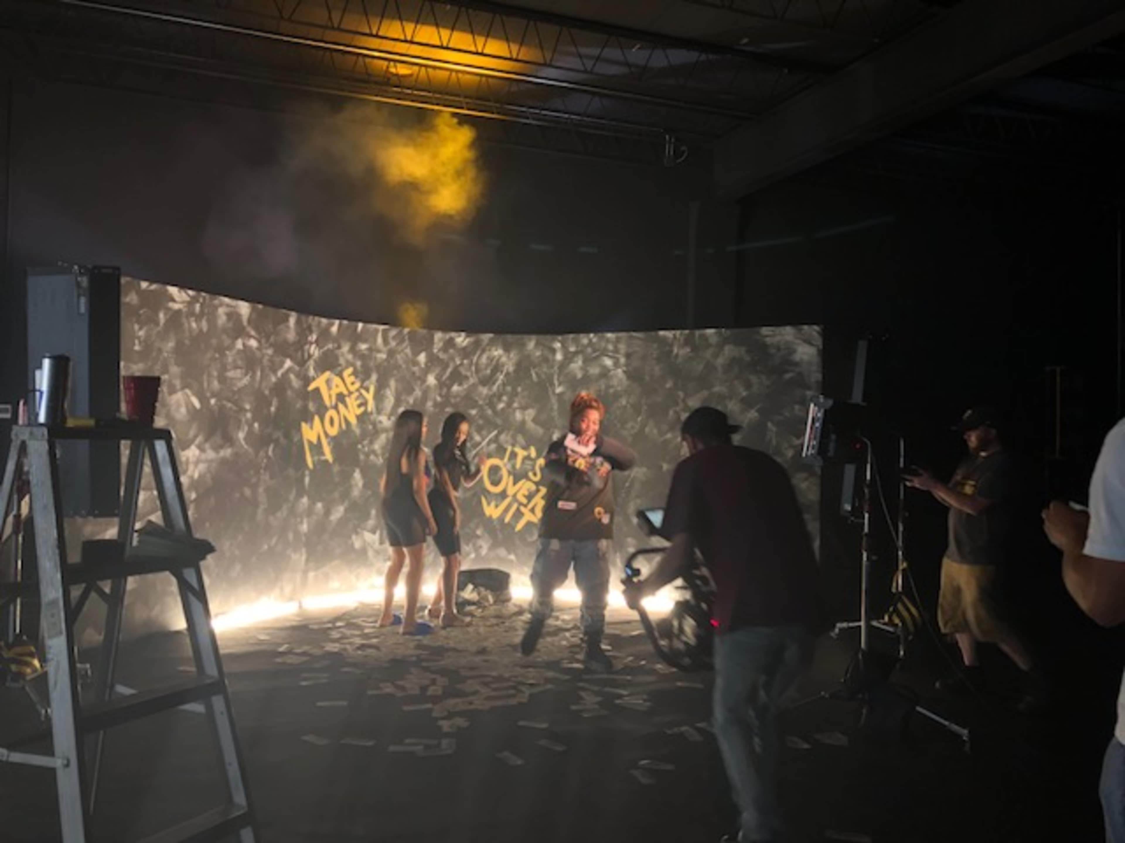 A film crew is shooting a scene with a backdrop of money and smoke, featuring a performer in front of two others in a studio setting.