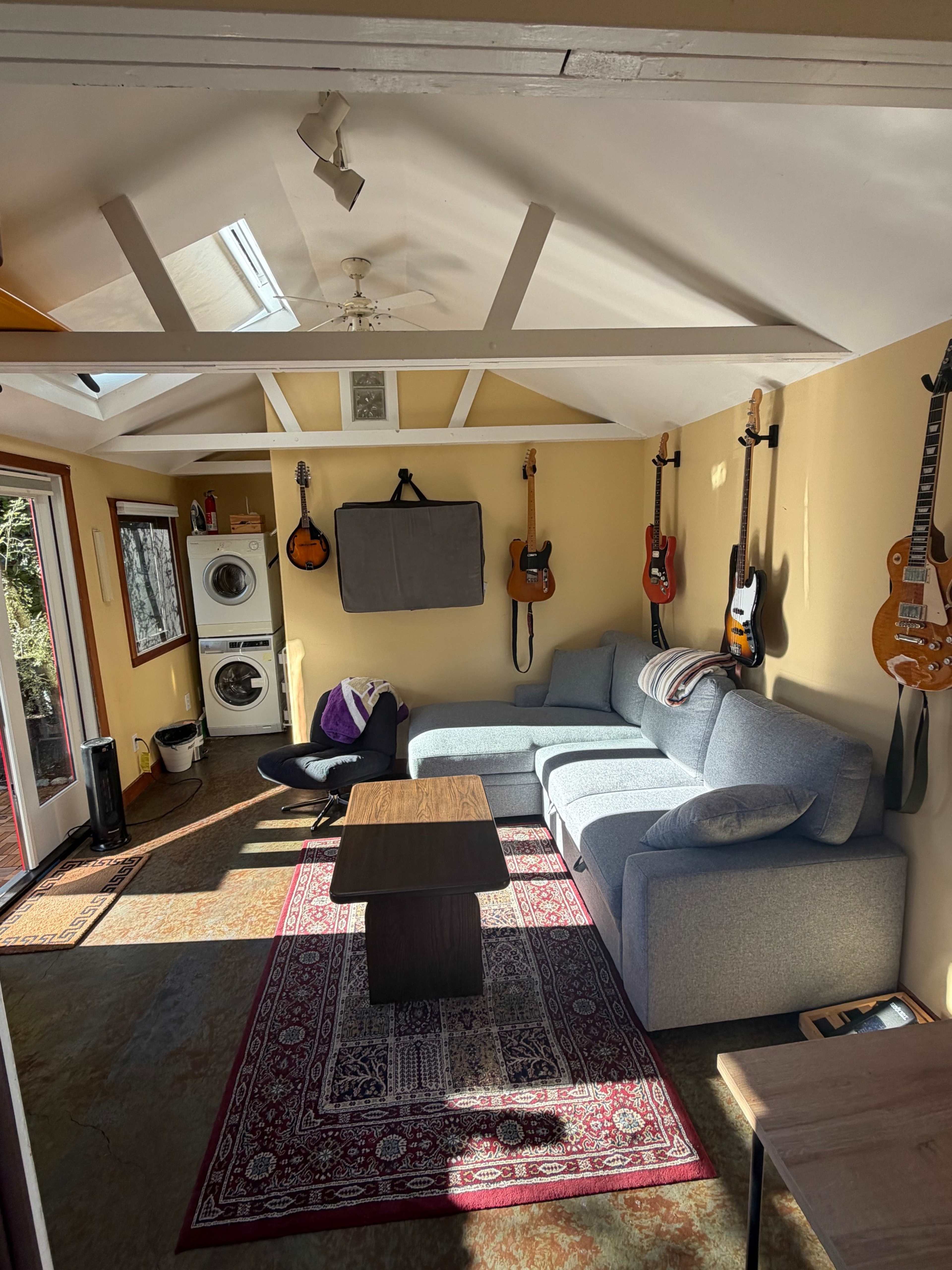 The image shows a cozy room with a gray sofa, a wooden coffee table, and several guitars hanging on the walls, along with a washing machine and a large window.