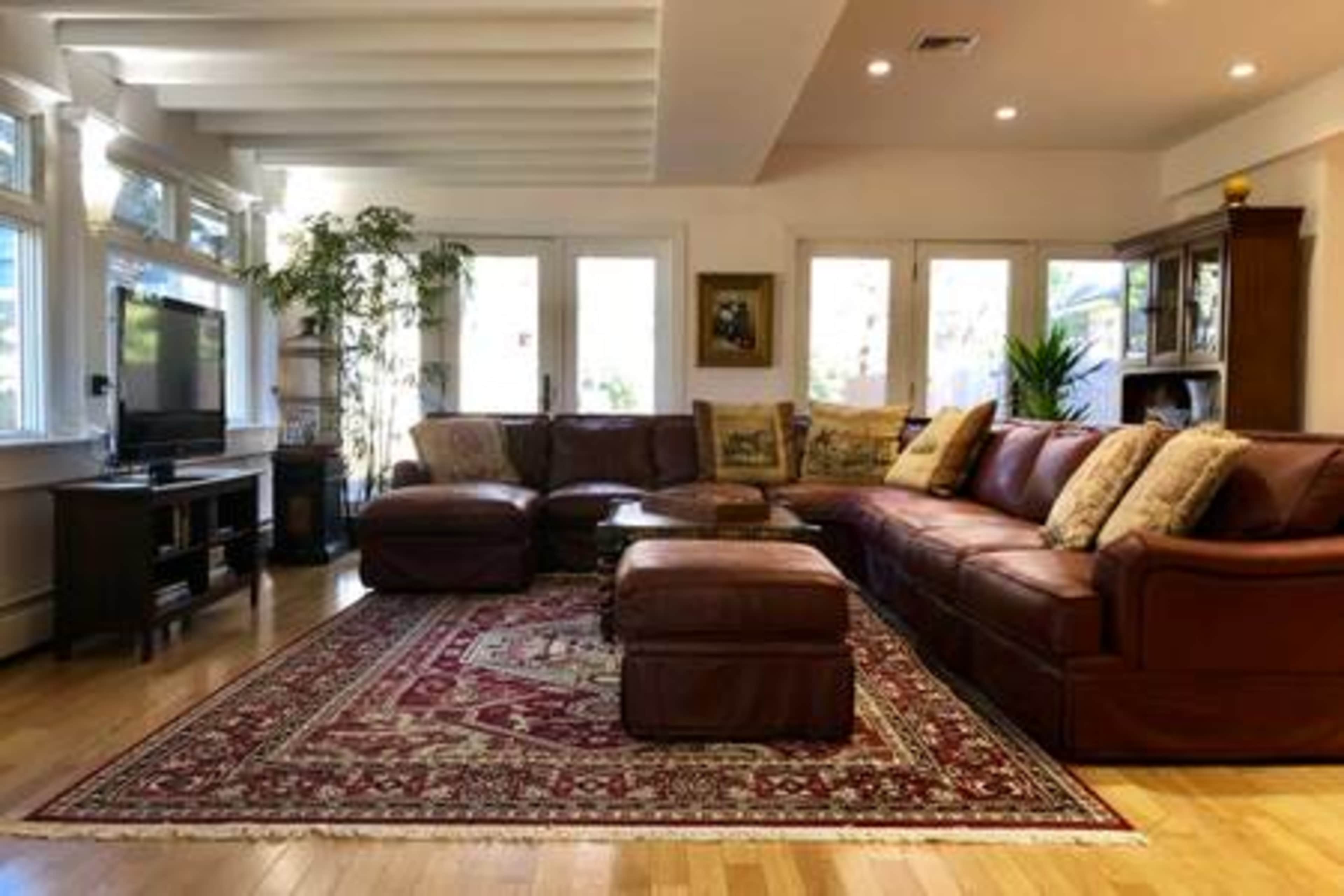 The image shows a bright living room featuring a brown leather sectional sofa, an area rug, and large windows with natural light.