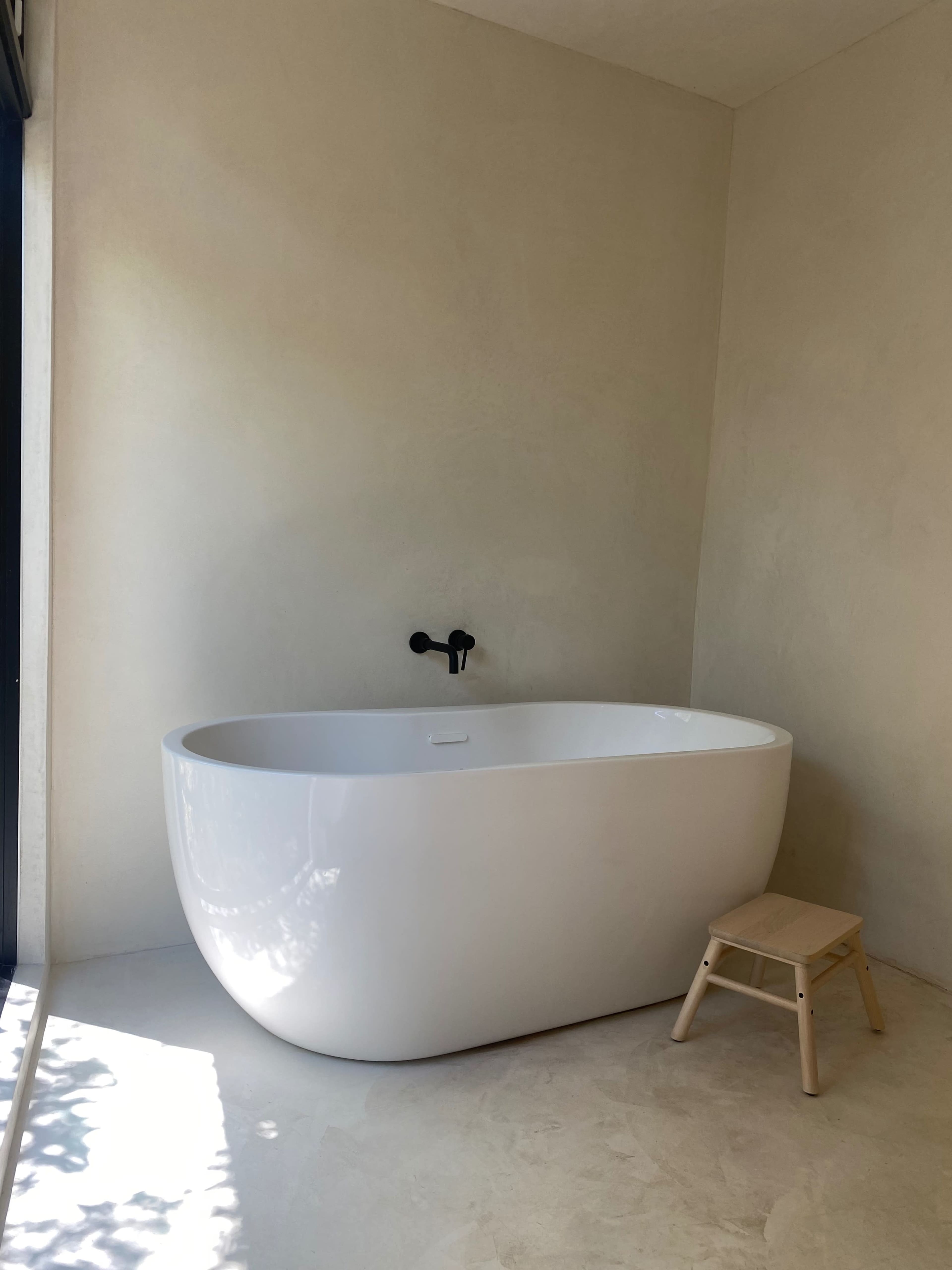 A modern, freestanding white bathtub beside a small wooden stool in a minimally designed bathroom with light-colored walls.