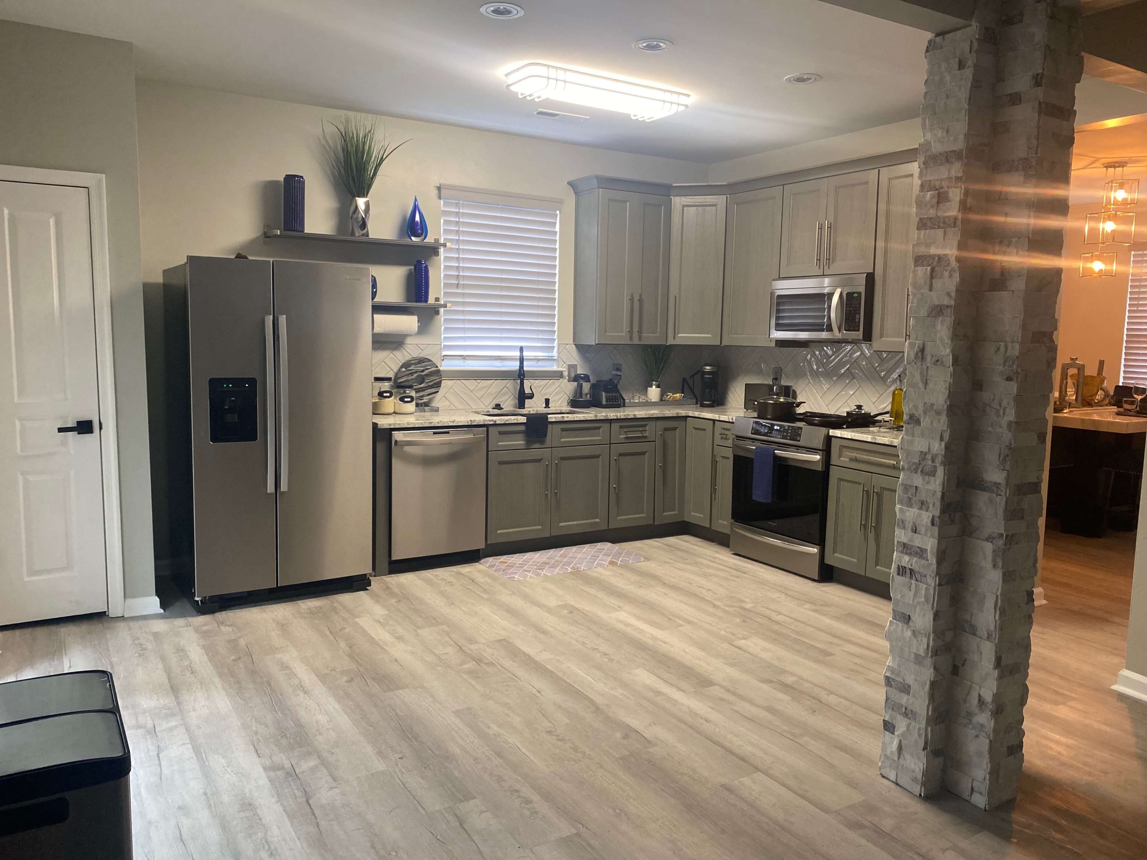 The image shows a modern kitchen with gray cabinets, stainless steel appliances, and a combination of tiled and wooden flooring.