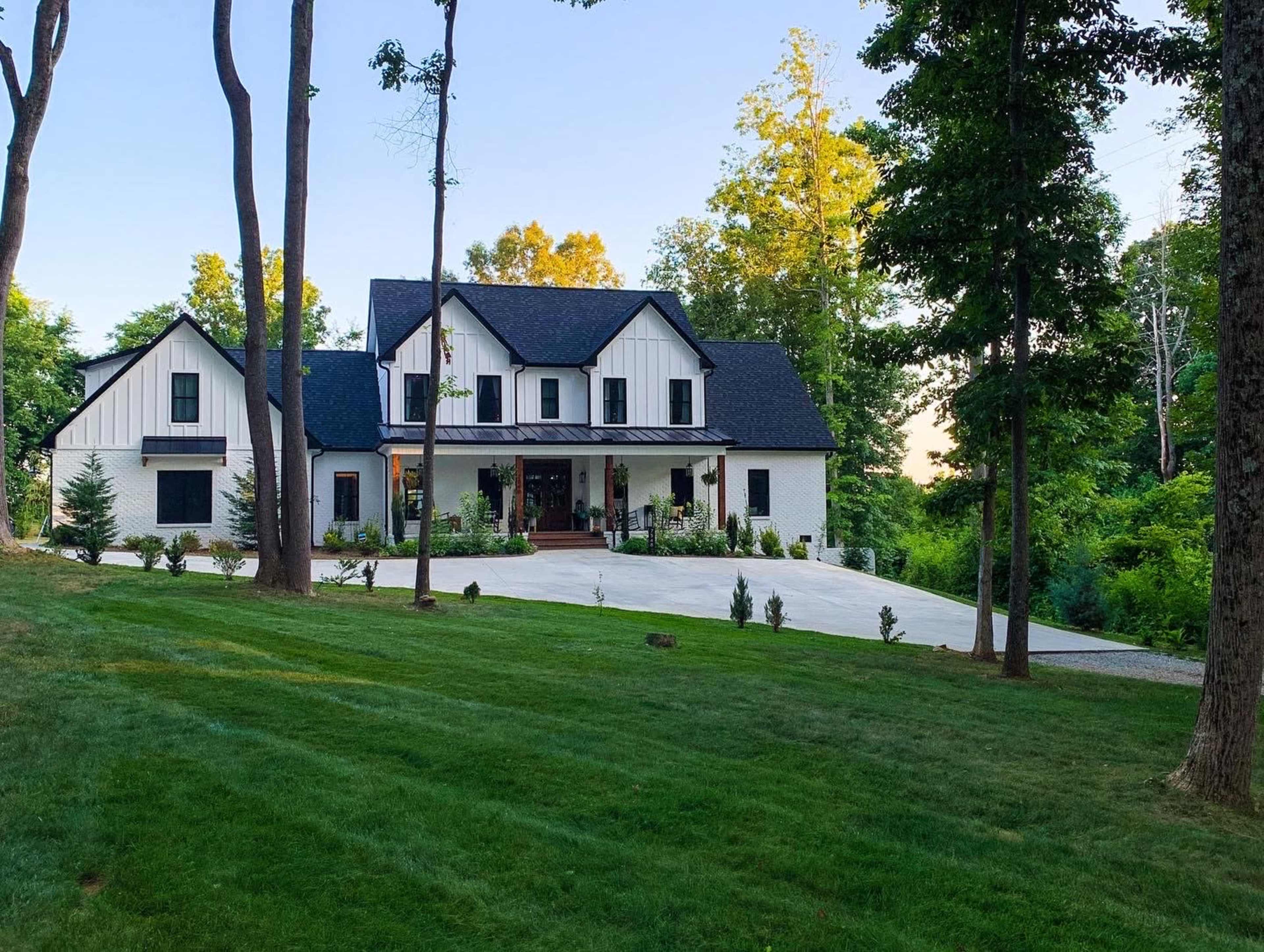 A modern two-story house with a black roof and white facade is situated on a green lawn surrounded by trees.