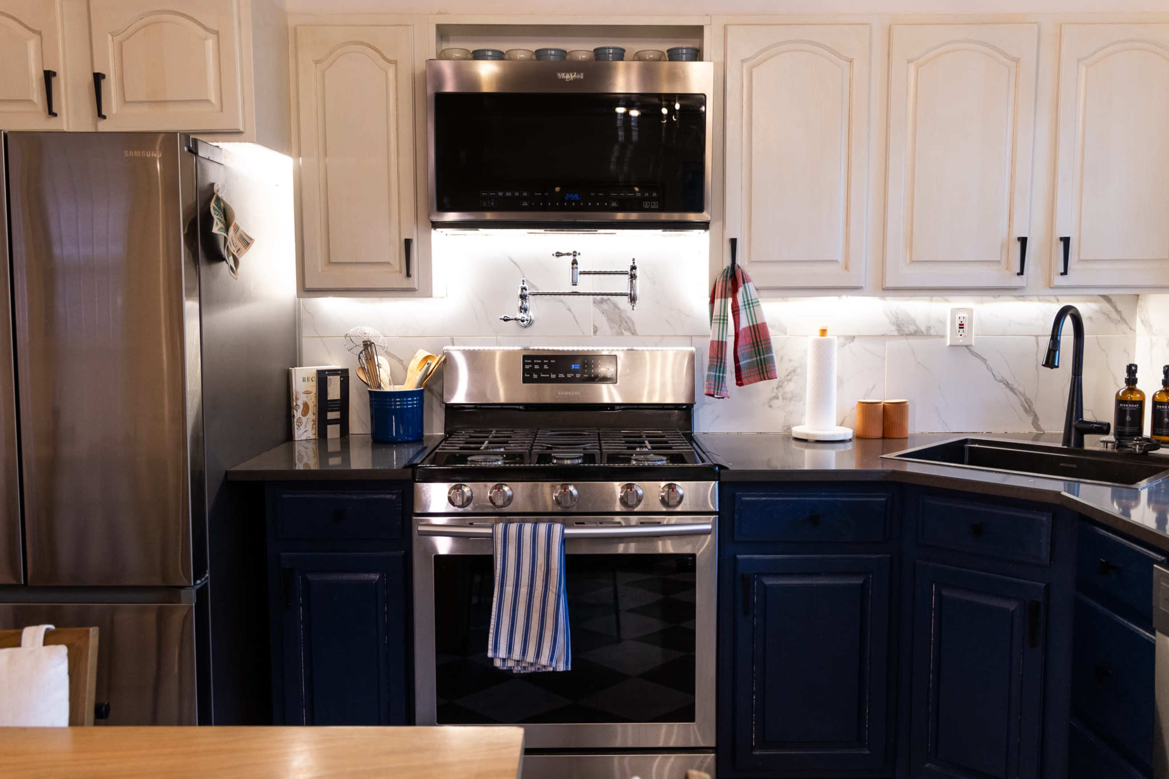 The image shows a kitchen featuring a stainless steel refrigerator, a gas stove with an oven, and dark blue cabinetry, alongside white countertops and a backsplash.