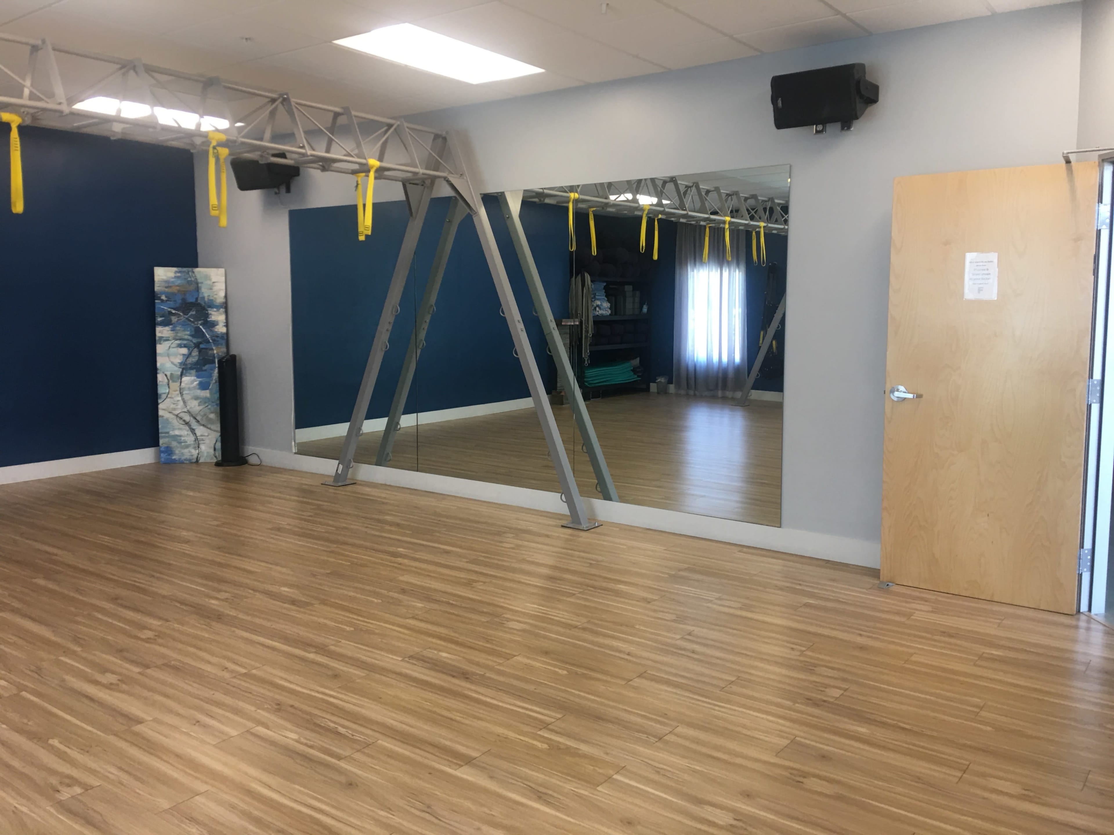 A spacious exercise room with a large mirror on one wall, wooden flooring, and fitness equipment suspended from the ceiling.
